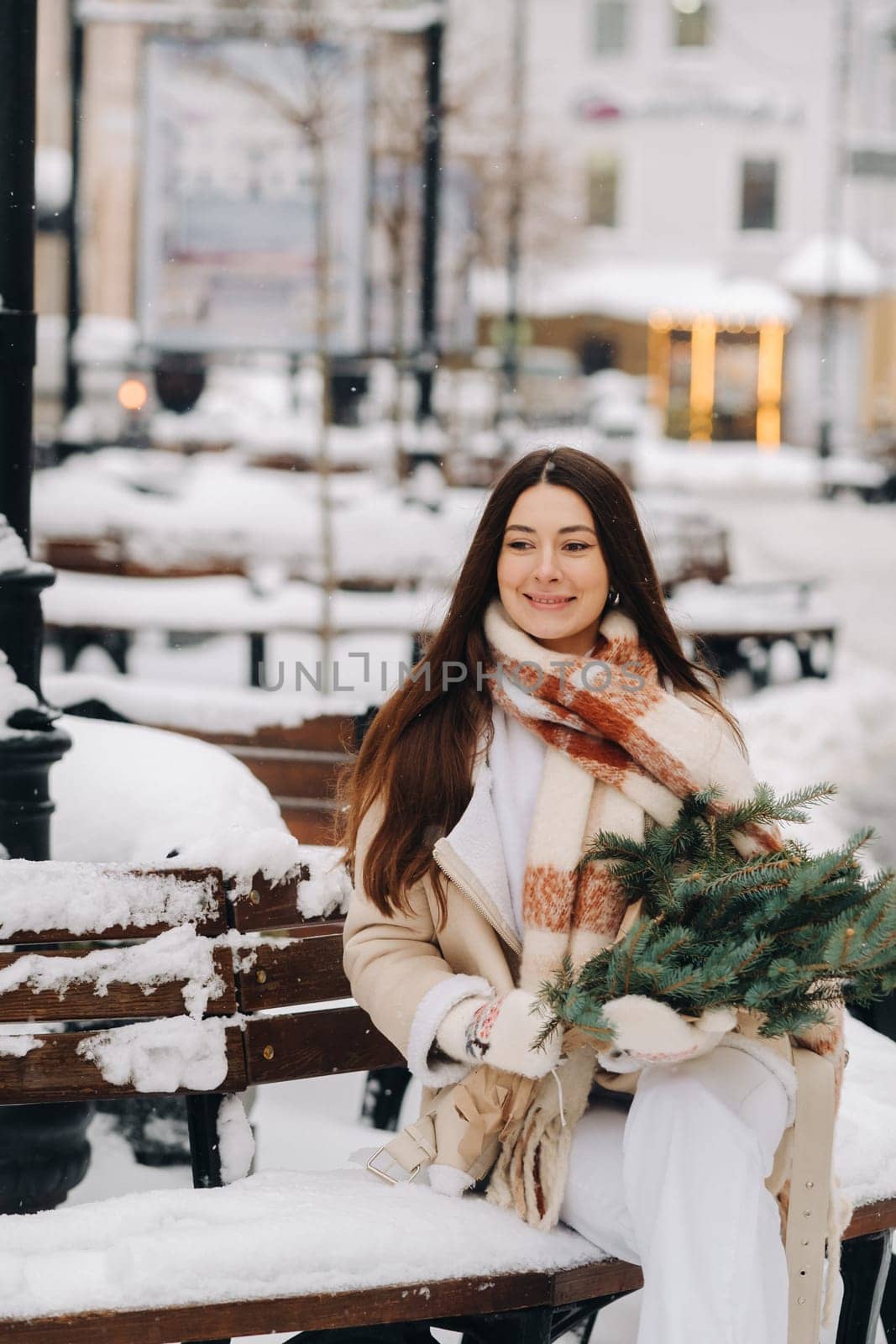 A girl with long hair in winter sits on a bench outside with a bouquet of fresh fir branches.