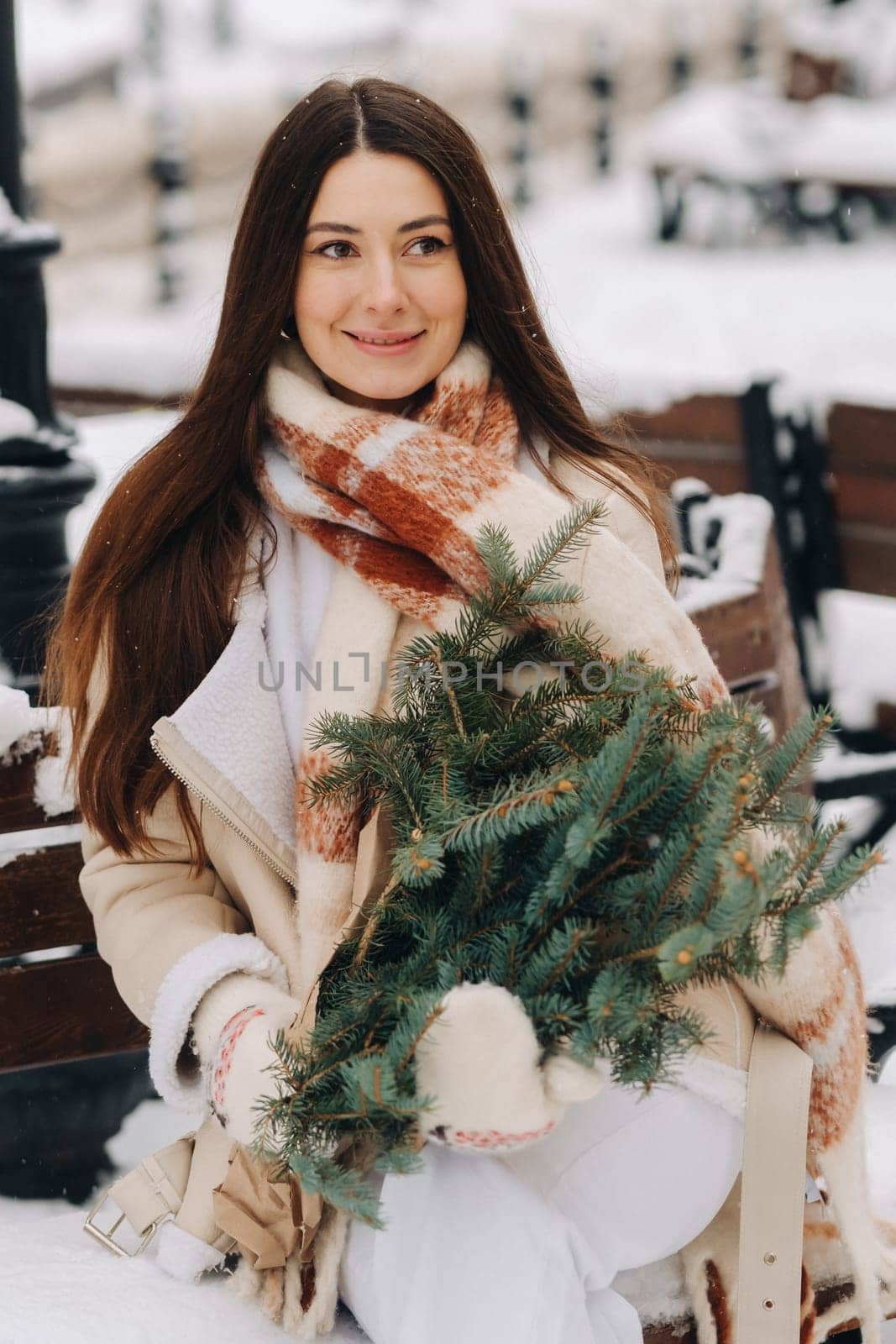 A girl with long hair in winter sits on a bench outside with a bouquet of fresh fir branches by Lobachad