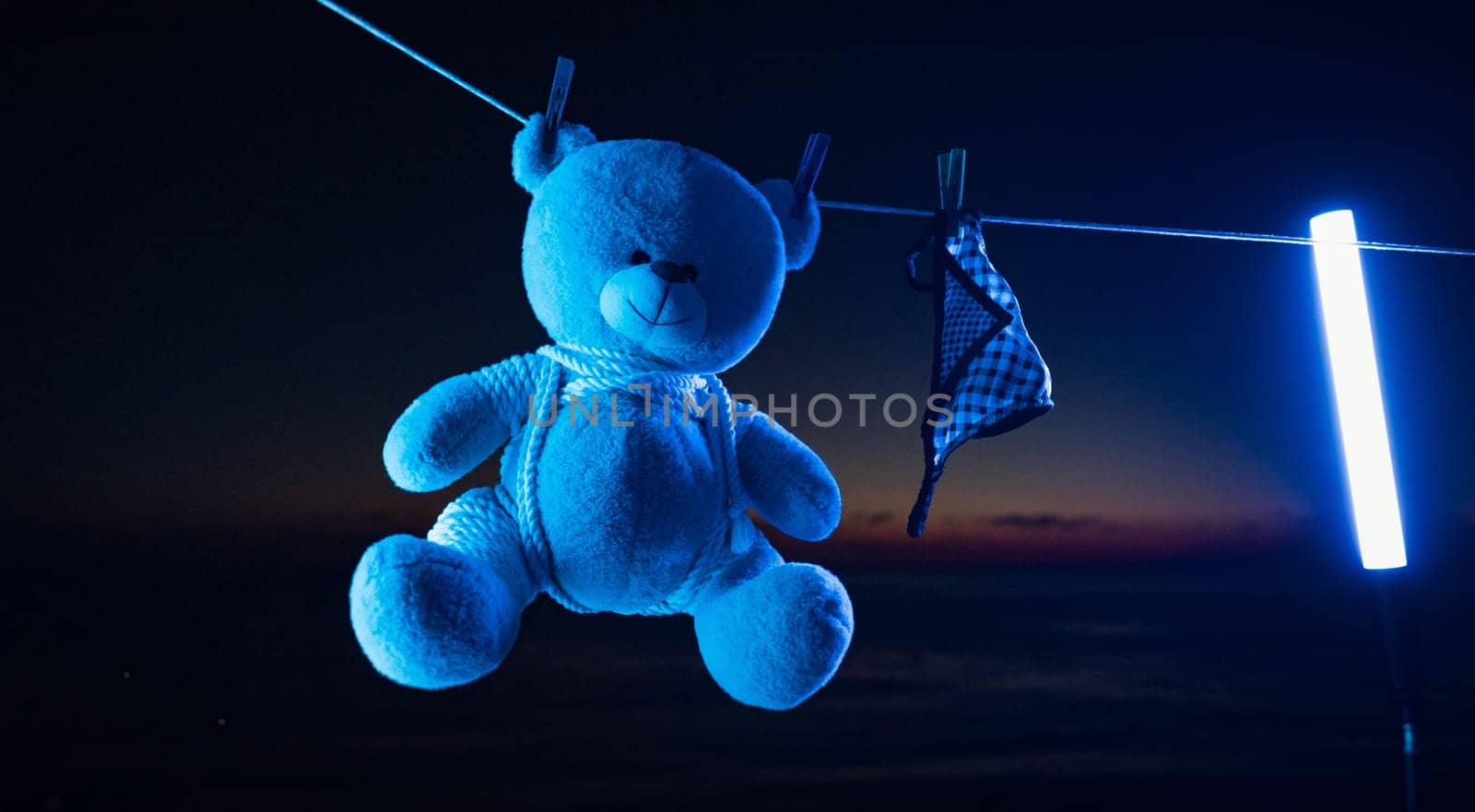 teddy bear toy hangs on a clothesline on clothespins in neon light. tied with shibari ropes