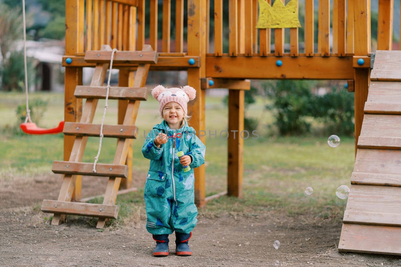 Little smiling girl stands with a bottle of soap bubbles near the climbing wall on the playground. High quality photo