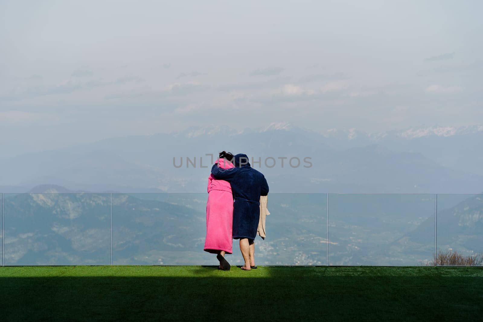 Back view of anonymous couple in robes looking away while standing closely and hugging on green lawn and admiring view of mountains and hazy nature in daylight