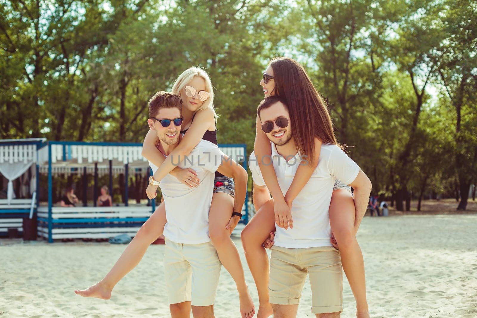 Group of friends walking along the beach, with men giving piggyback ride to girlfriends. Happy young friends enjoying a day at beach.