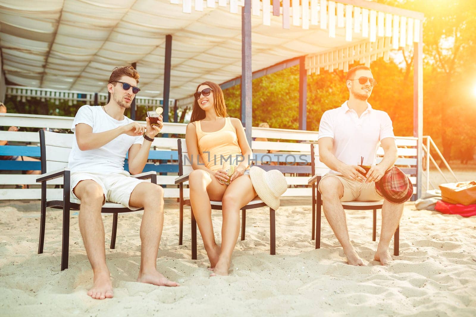 Lifestyle Young people enjoying summer vacation sunbathing drinking at beach bar. Two guys and a young girl sit on the terrace of the summer bar and enjoy the conversation Sun flare