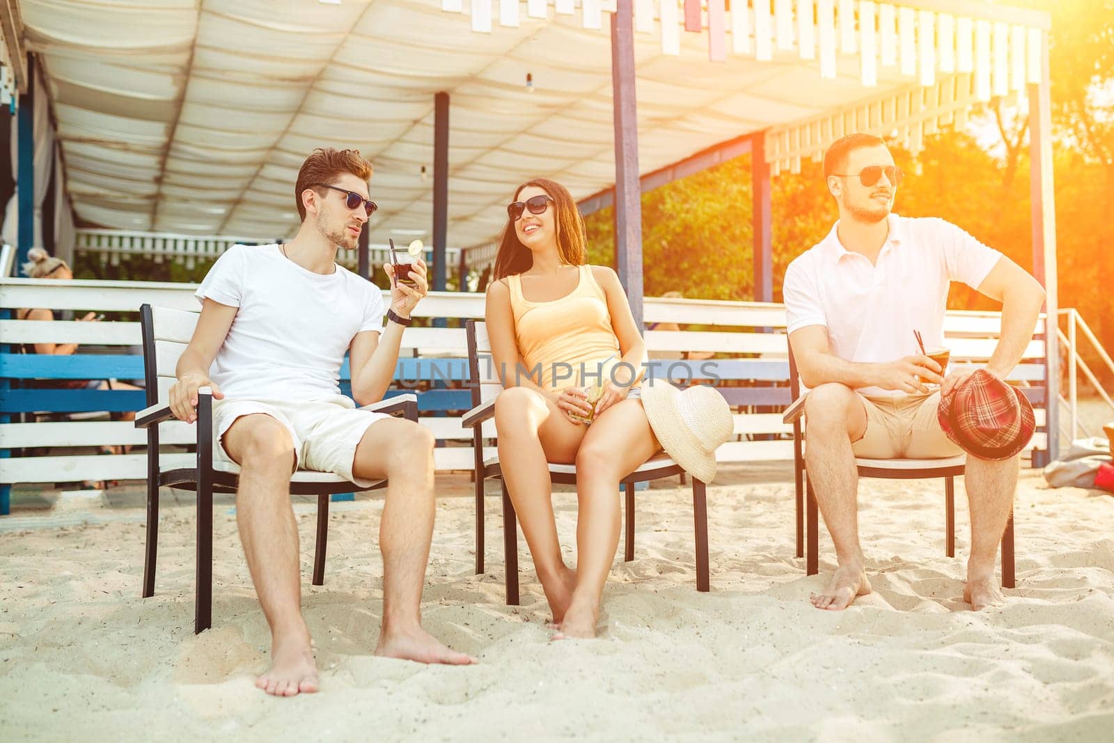 Lifestyle Young people enjoying summer vacation sunbathing drinking at beach bar. Two guys and a young girl sit on the terrace of the summer bar and enjoy the conversation Sun flare