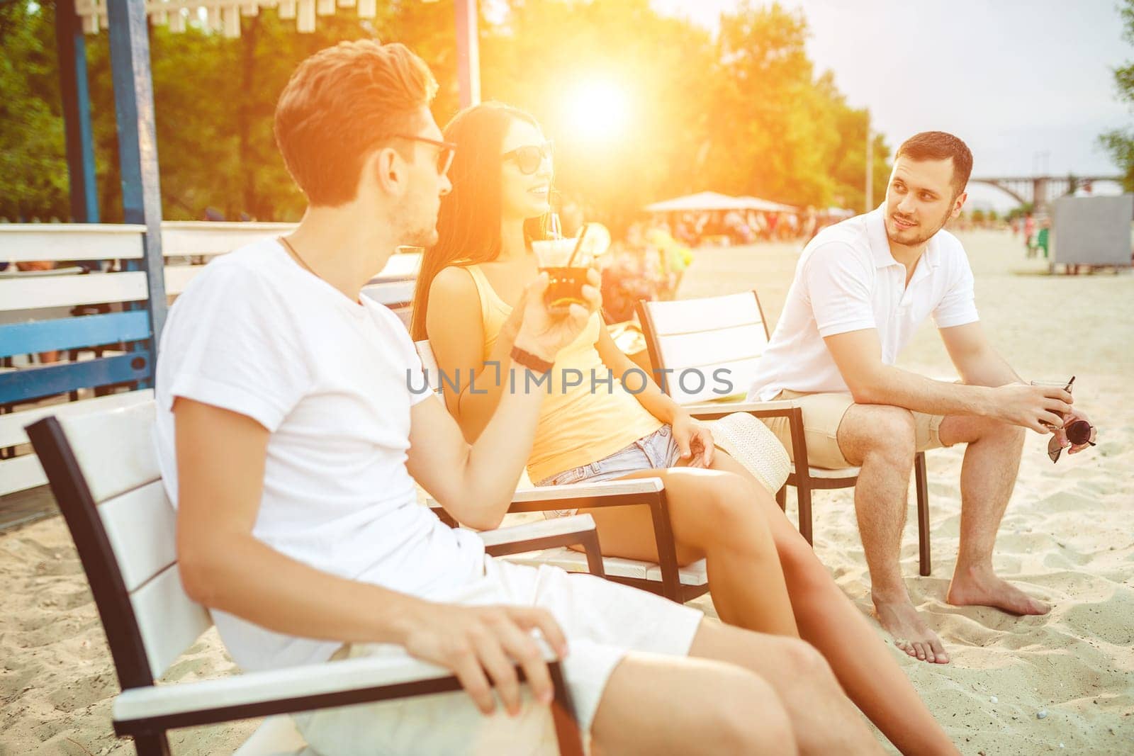 Lifestyle Young people enjoying summer vacation sunbathing drinking at beach bar. Two guys and a young girl sit on the terrace of the summer bar and enjoy the conversation Sun flare