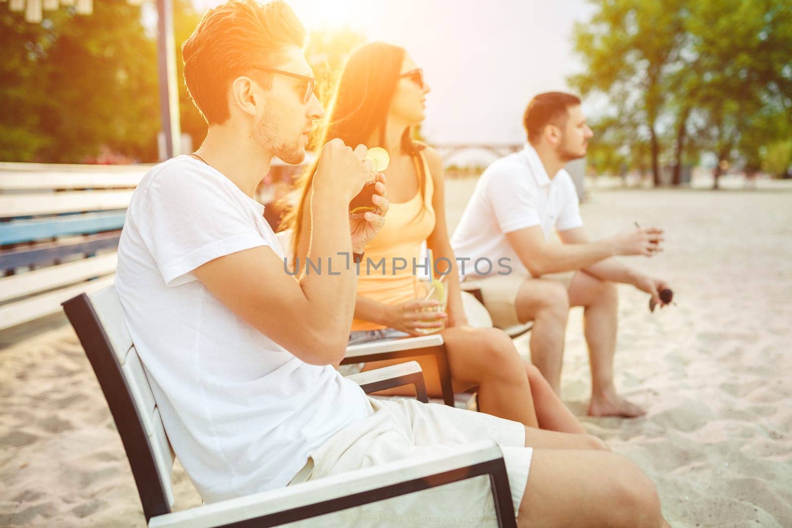 Lifestyle Young people enjoying summer vacation sunbathing drinking at beach bar. Two guys and a young girl sit on the terrace of the summer bar and enjoy the conversation Sun flare