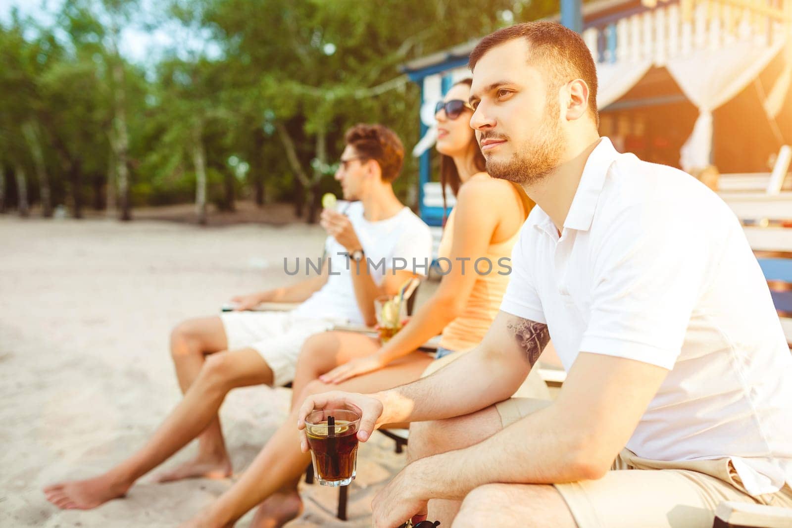 Lifestyle Young people enjoying summer vacation sunbathing drinking at beach bar. Two guys and a young girl sit on the terrace of the summer bar and enjoy the conversation Sun flare