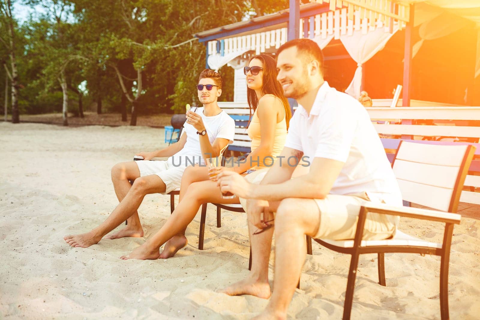 Lifestyle Young people enjoying summer vacation sunbathing drinking at beach bar. Two guys and a young girl sit on the terrace of the summer bar and enjoy the conversation Sun flare