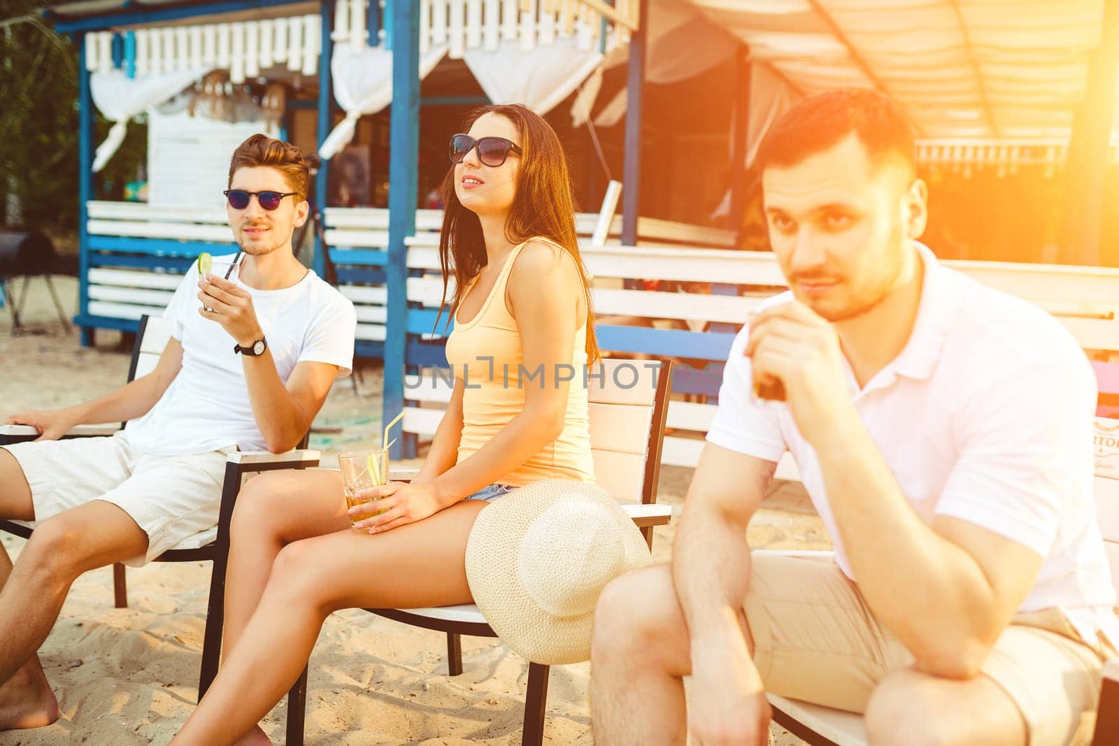 Lifestyle Young people enjoying summer vacation sunbathing drinking at beach bar. Two guys and a young girl sit on the terrace of the summer bar and enjoy the conversation Sun flare