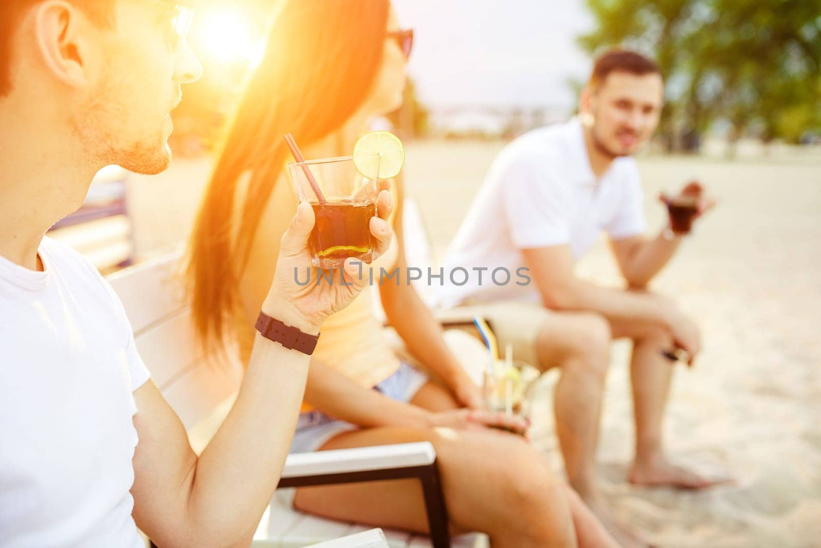 Lifestyle Young people enjoying summer vacation sunbathing drinking at beach bar. Two guys and a young girl sit on the terrace of the summer bar and enjoy the conversation Sun flare