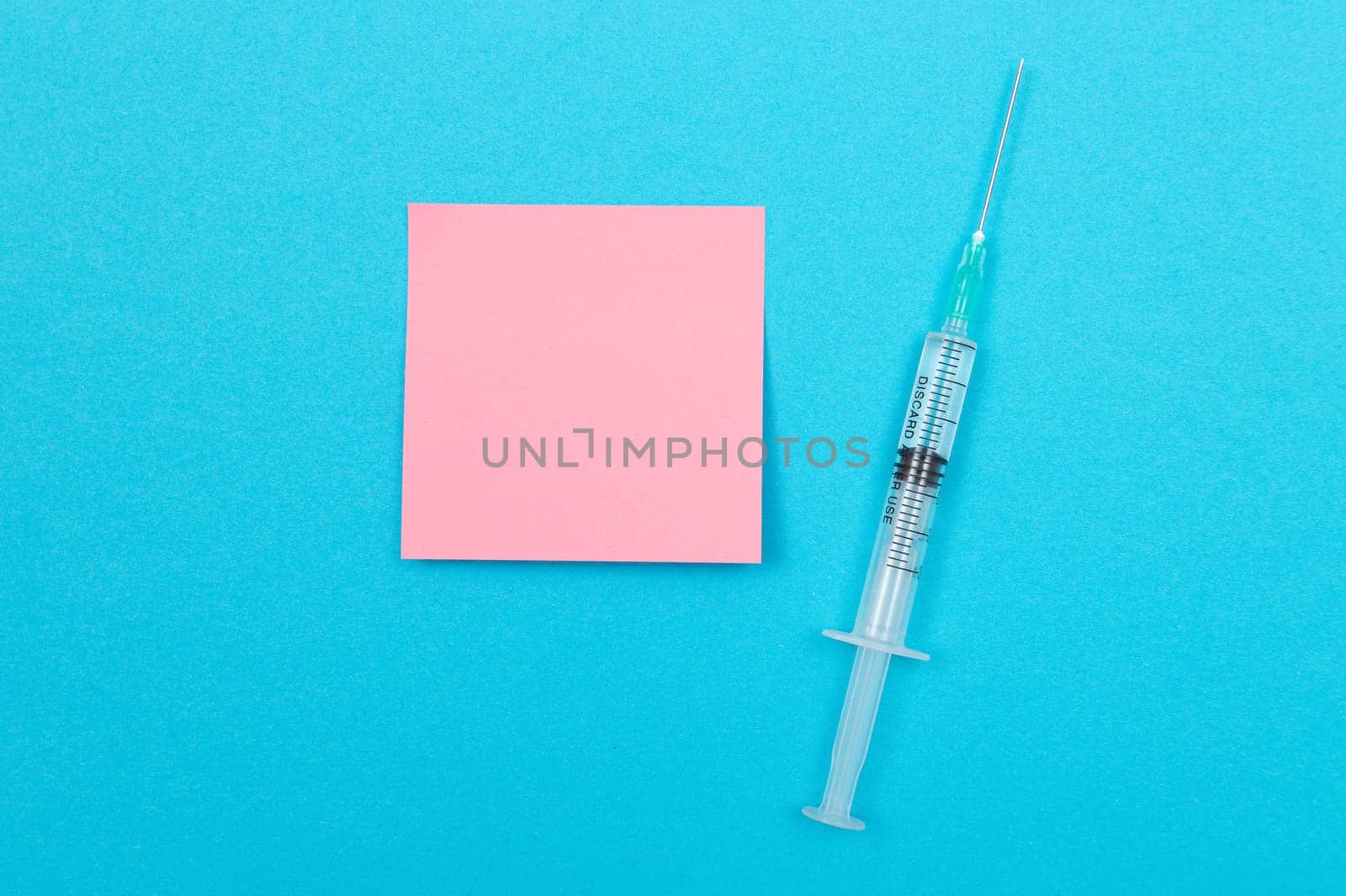 Vaccination, Immunology or Revaccination Concept - A Medical Disposable Syringe Lying on Blue Table in Doctor's Office in a Hospital or Clinic. Blank Pink Sticky Note - Mock Up with Copy Space