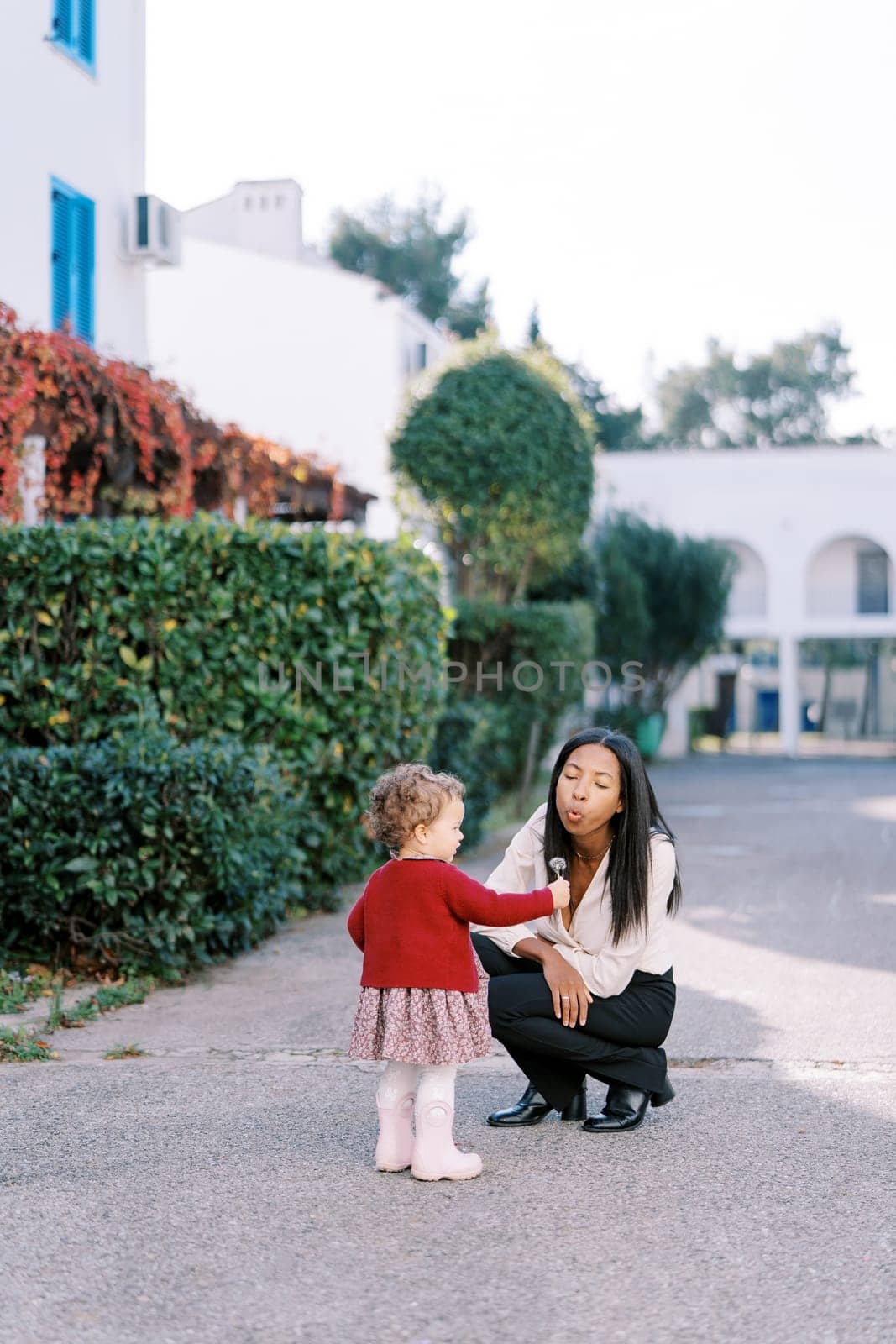 Little girl holds out a dandelion to her mother squatted on the road near the house. High quality photo