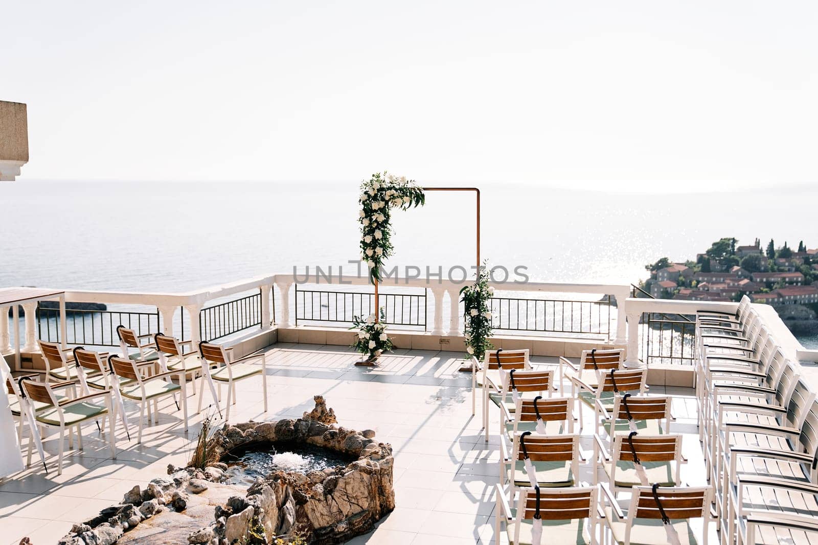Rows of chairs stand near a fountain in front of a wedding arch on a terrace above the island of Sveti Stefan. Montenegro. High quality photo