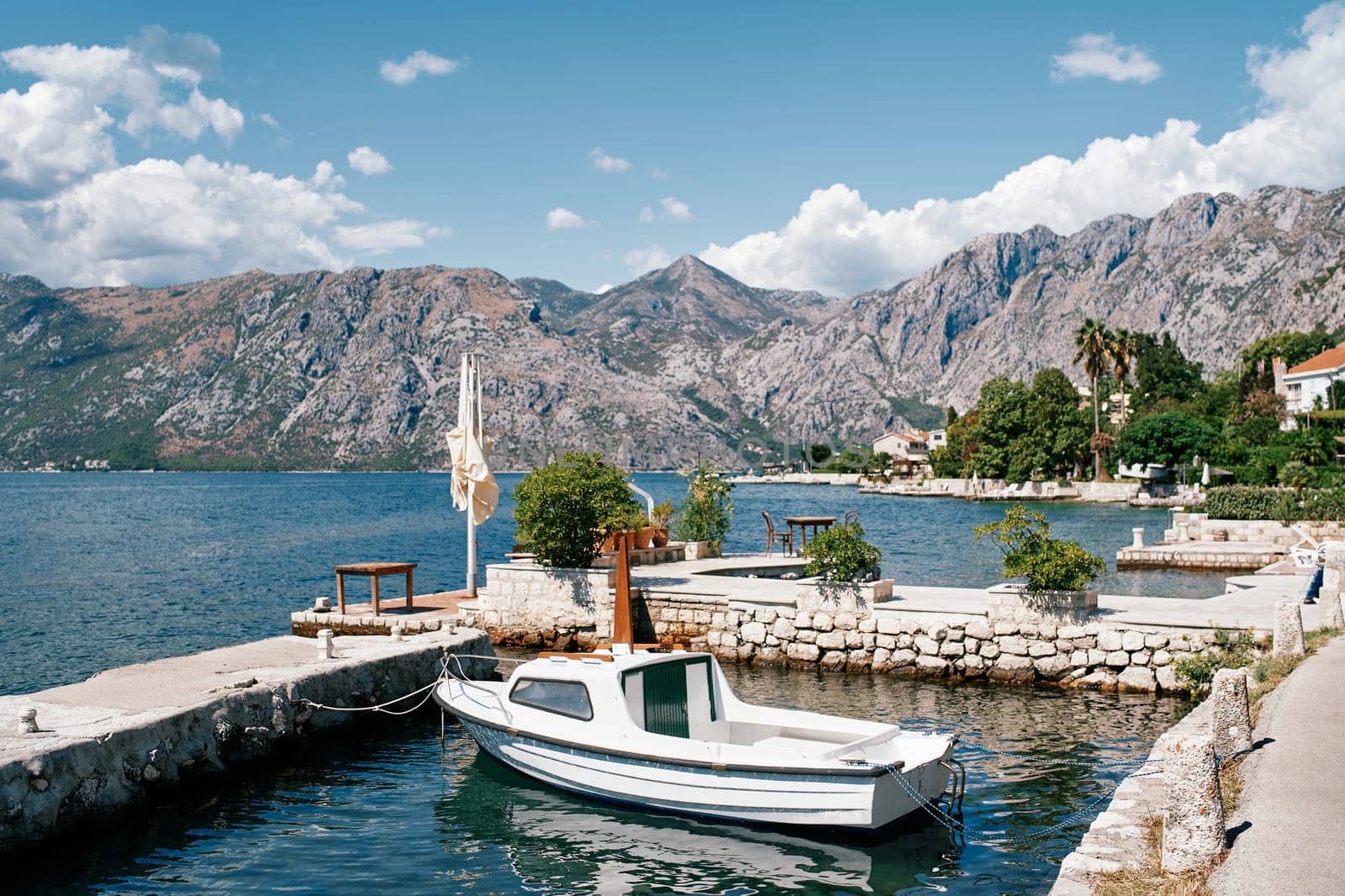 Small boat is moored at the stone pier of an ancient town at the foot of the mountains. Dobrota, Montenegro by Nadtochiy