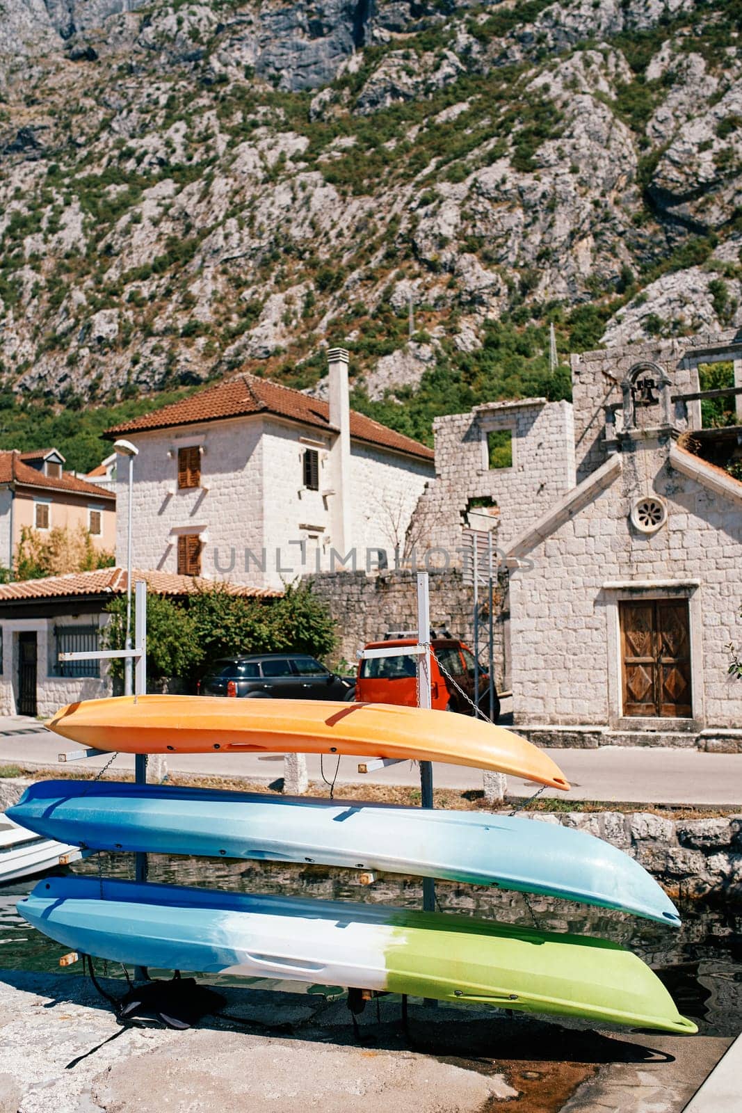Multi-colored kayaks dry upside down on a rack near old houses at the foot of the mountains by Nadtochiy