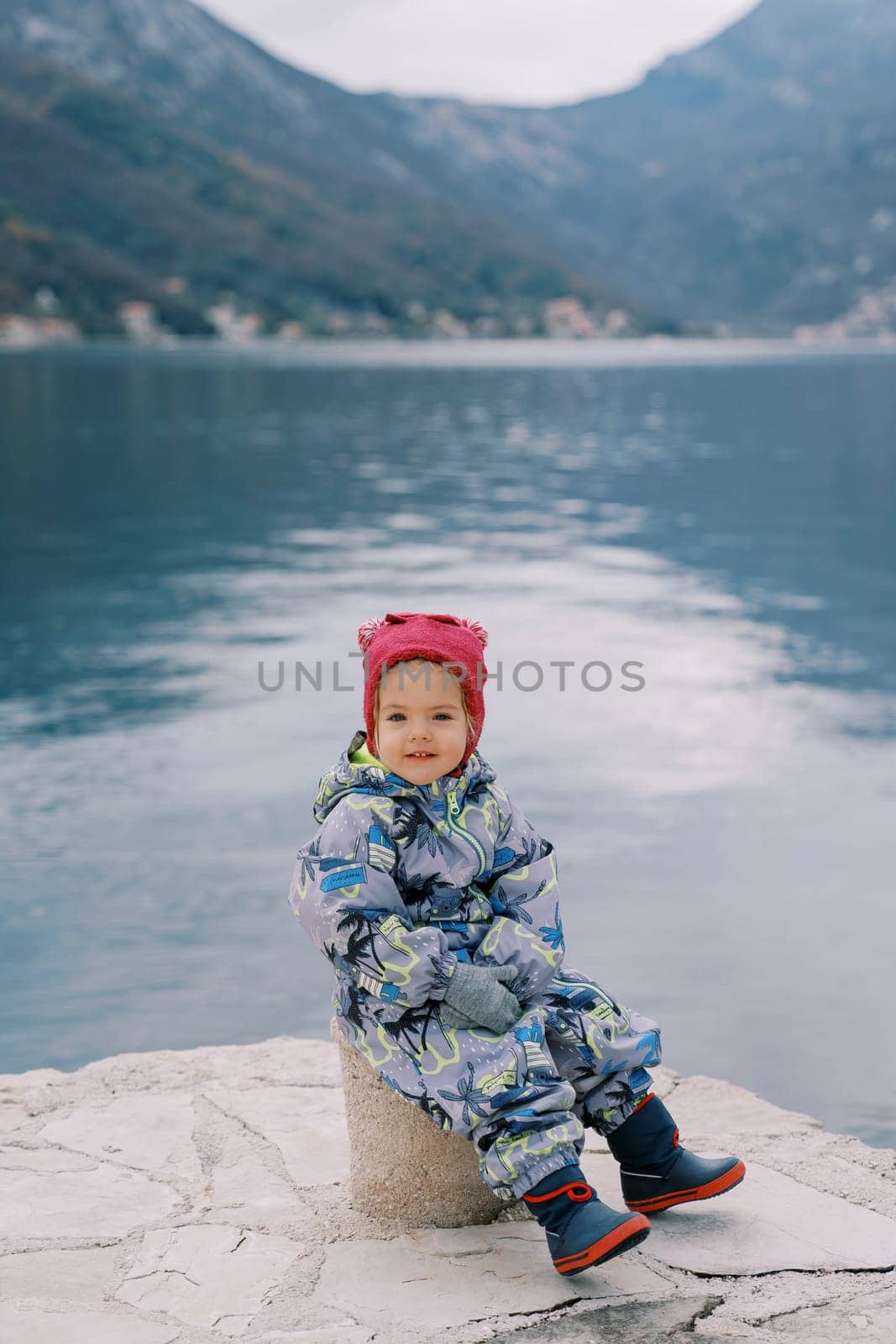Little smiling girl sitting on a bollard on the pier. High quality photo