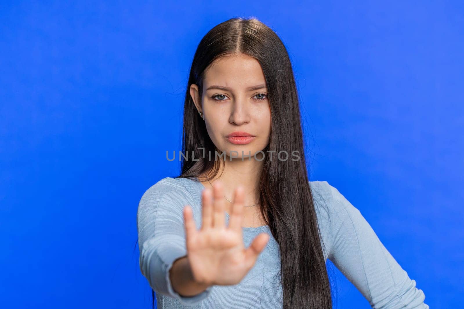 Come here, join us. Welcome. Brunette attractive woman showing inviting gesture with hands, ask to join, beckoning to coming, gesturing hello, goodbye. Strict girl isolated on blue studio background