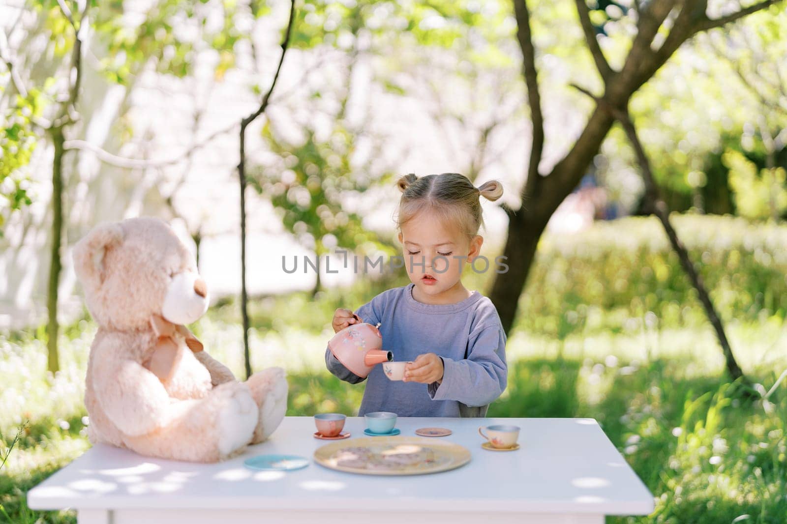 Little girl pours tea from a toy teapot into a toy cup while playing with a teddy bear in a tea party. High quality photo
