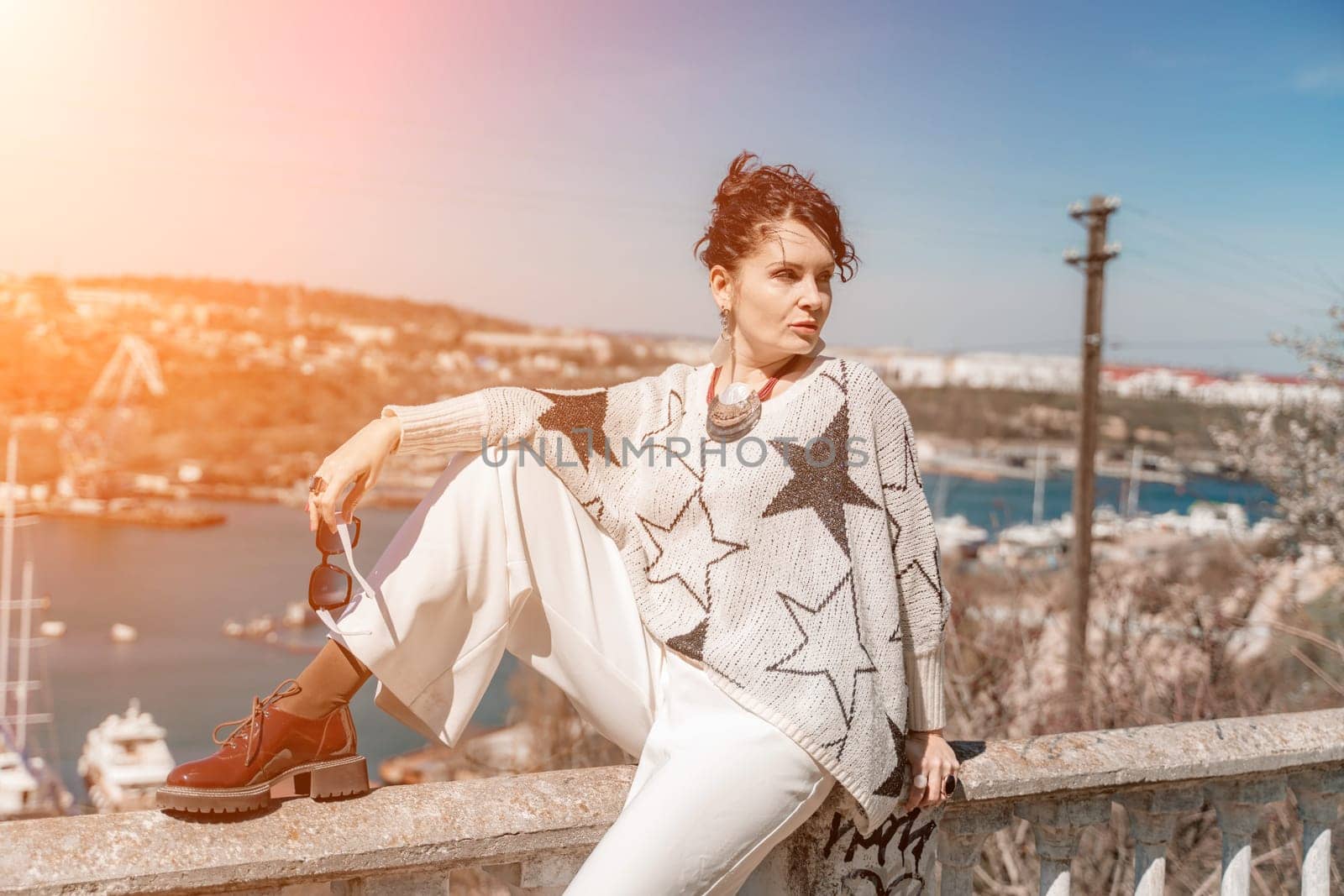 Woman walks around the city, lifestyle. A young beautiful woman in white trousers and a sweater sits on a white fence with balusters and overlooks the sea bay and the city. by Matiunina