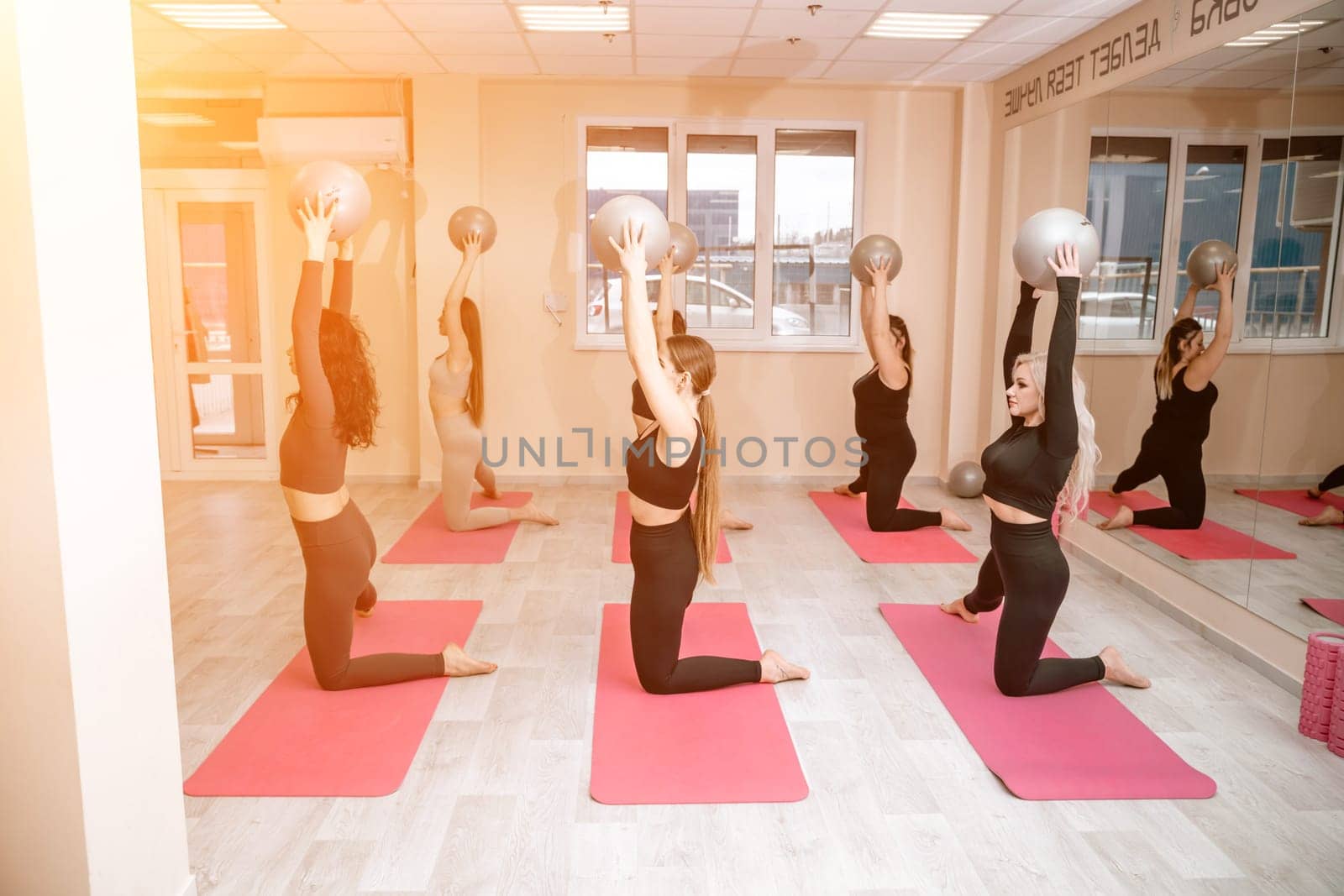 A group of six athletic women doing pilates or yoga on pink mats in front of a window in a beige loft studio interior. Teamwork, good mood and healthy lifestyle concept. by Matiunina