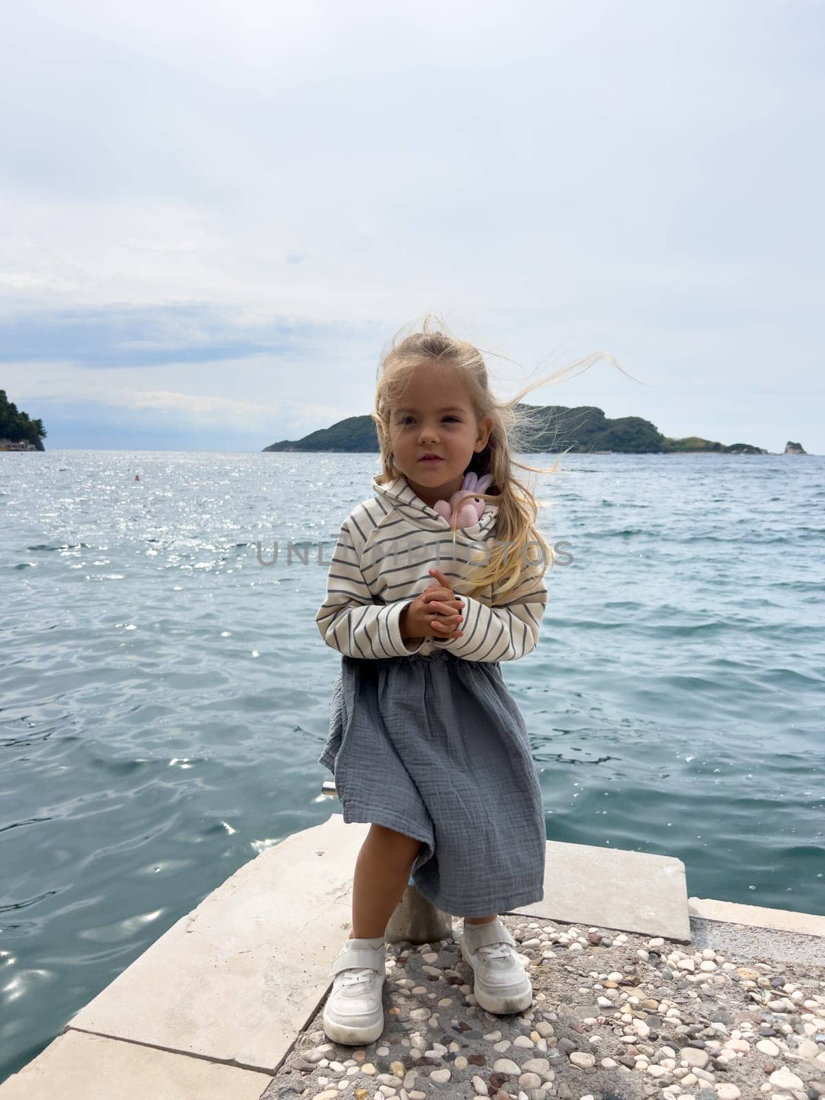 Little girl with flying hair sits on a bollard on a pier by the sea by Nadtochiy