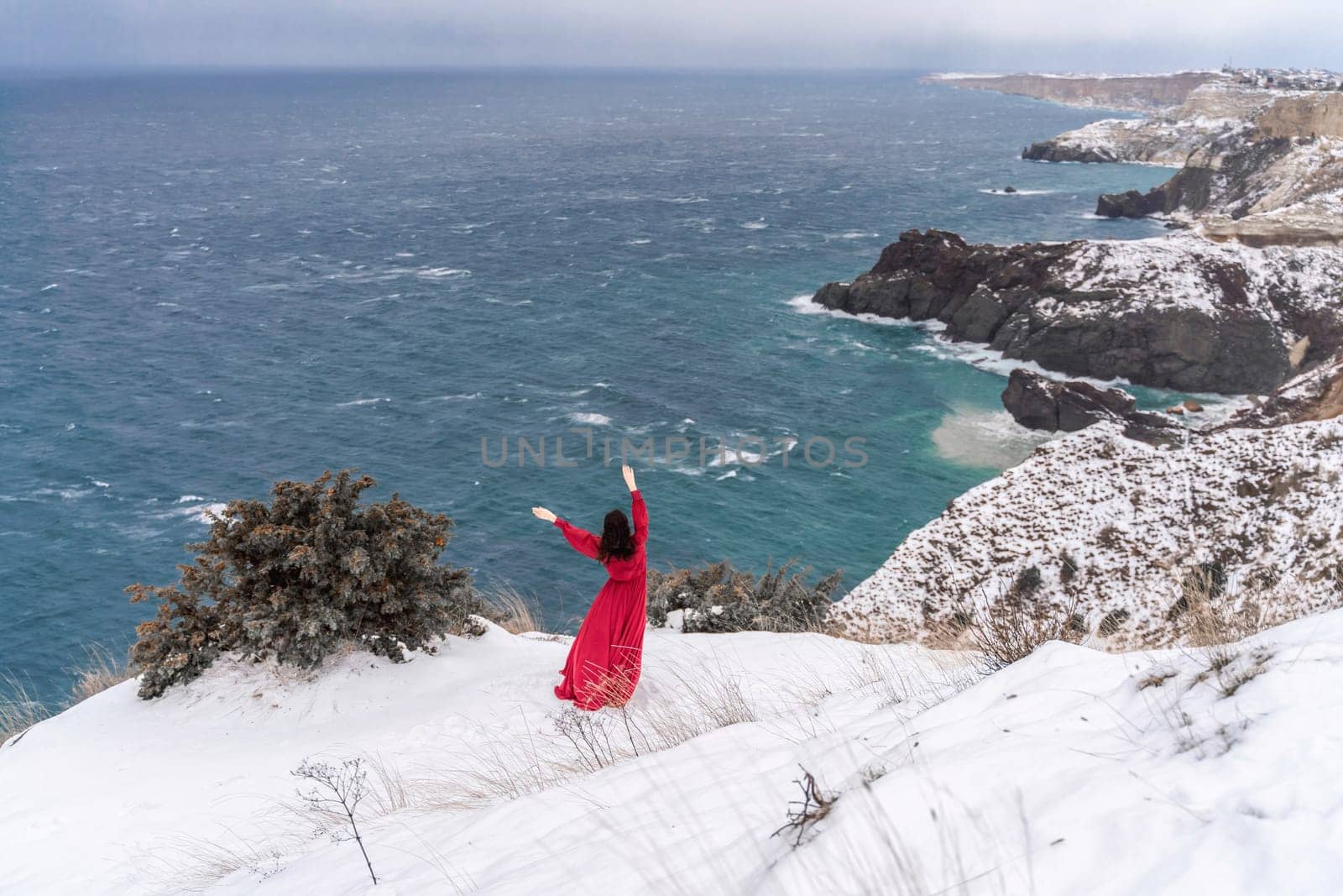 Woman red dress snow sea. Happy woman in a red dress in the snowy mountains by the emerald sea. The wind blows her clothes, posing against sea and snow background. by Matiunina