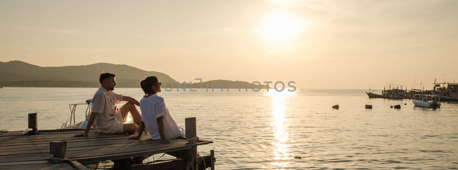 A couple of European men and an Asian woman on vacation in Thailand, couple watching the sunset sitting on a wooden deck pier in the ocean in Samaesan Thailand