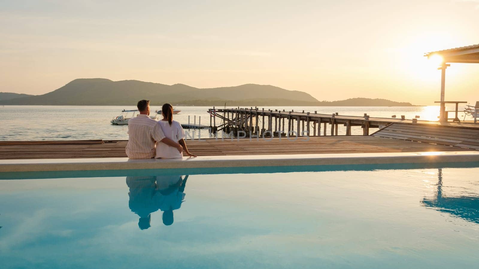 A couple of European men and an Asian woman on vacation in Thailand sitting on the edge of a pool by the ocean, Wooden pier in the ocean during sunset in Samaesan Thailand