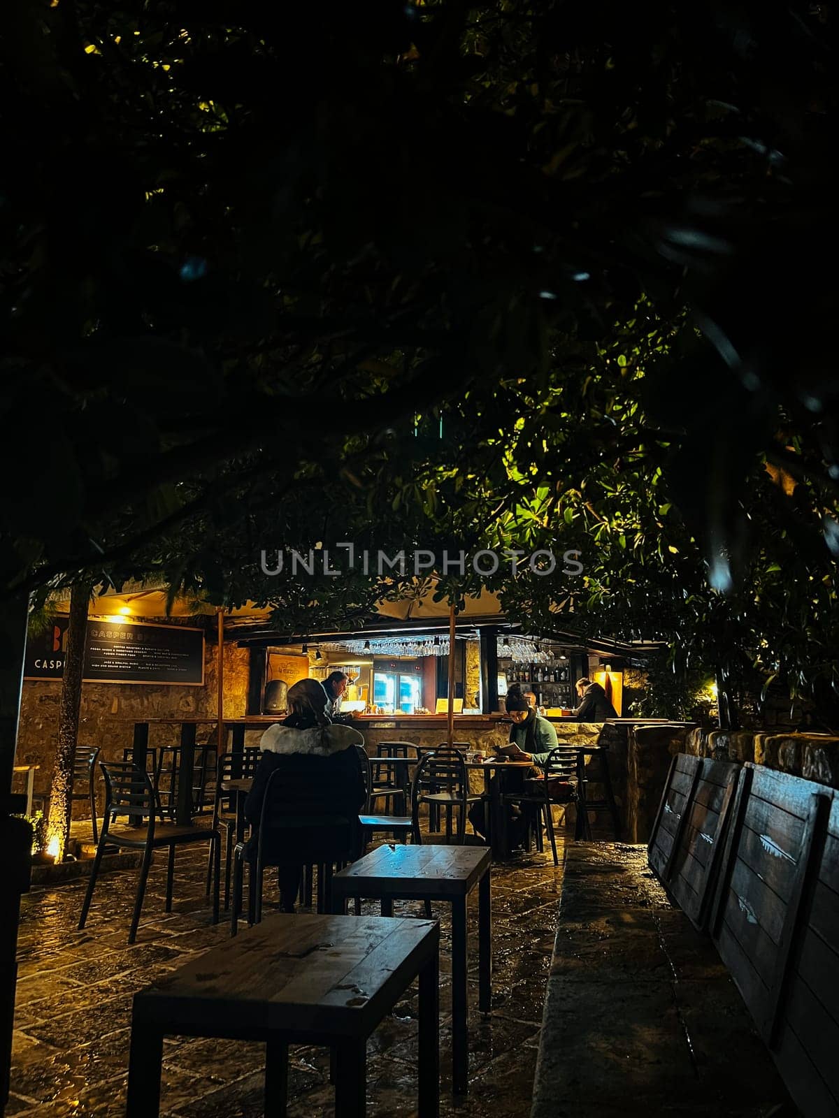 Budva, Montenegro - 25 december 2022: People sit on the chairs of a street cafe in the light of lanterns at night by Nadtochiy