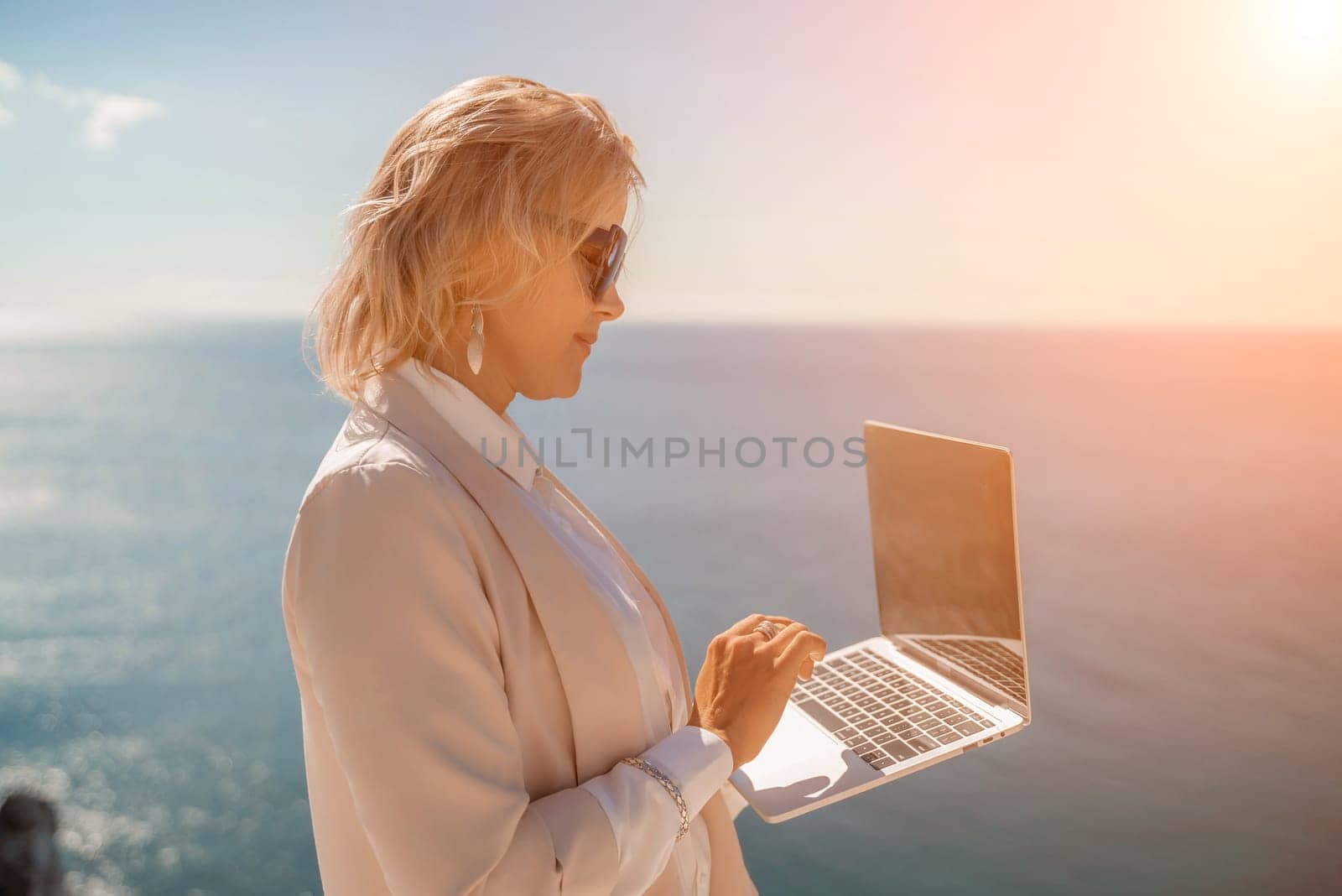 Freelance women sea working on the computer. Good looking middle aged woman typing on a laptop keyboard outdoors with a beautiful sea view. The concept of remote work