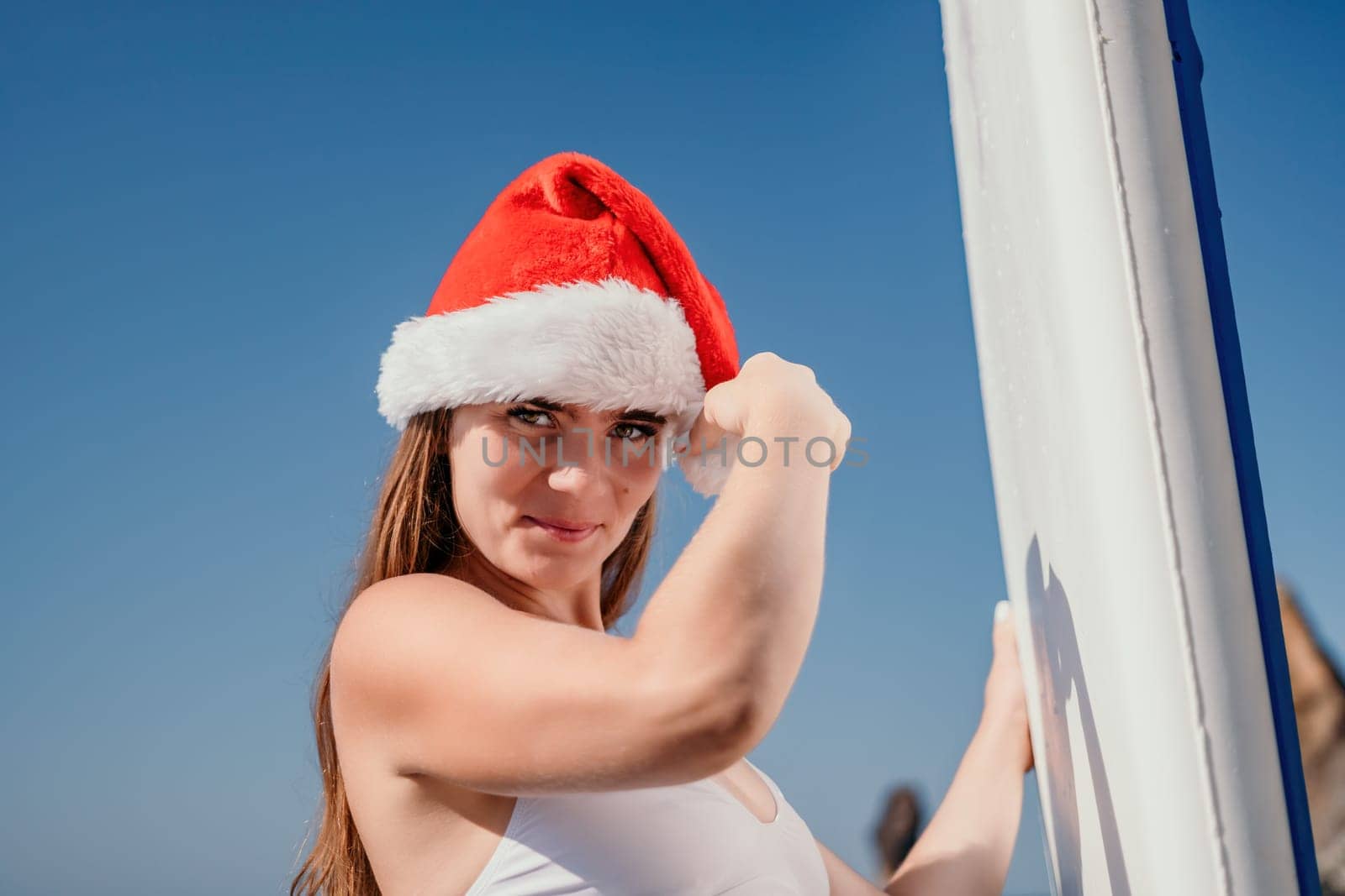 Woman sea sup. Close up portrait of happy young caucasian woman with long hair in Santa hat looking at camera and smiling. Cute woman portrait in a white bikini posing on sup board in the sea by panophotograph