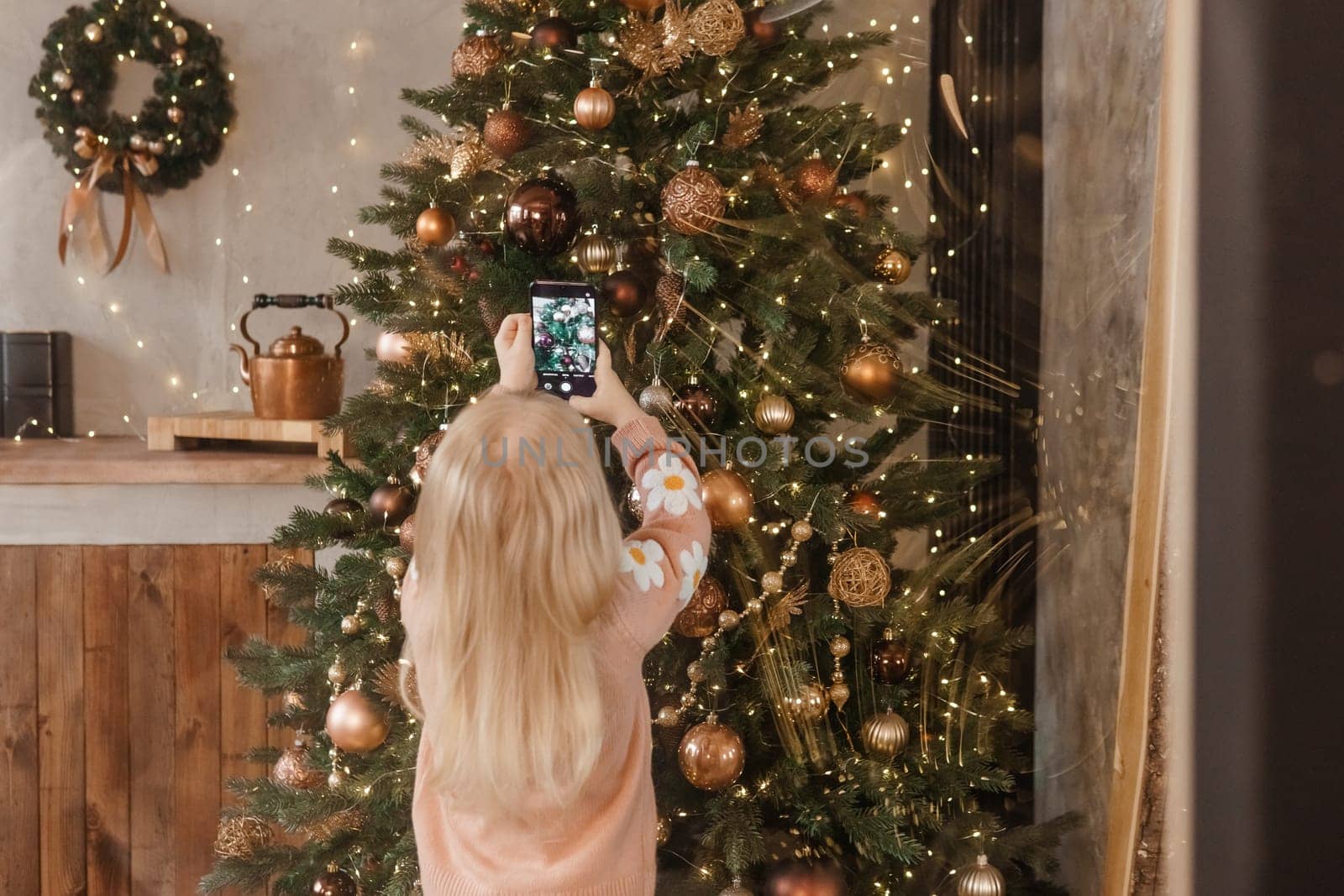 A little blonde girl photographs balloons on a Christmas tree in a festive interior decorated in a New Year's style. The concept of a merry Christmas