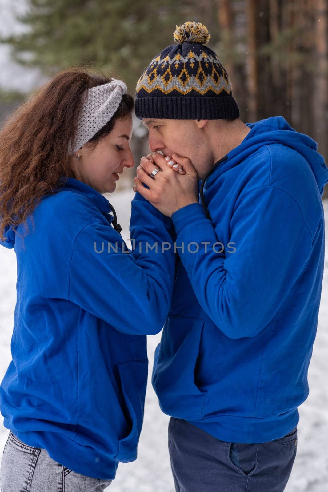 Outdoor happy couple in love posing in cold winter weather. A man and a woman in blue hoodies. Emotional young couple having fun while walking by winter forest, loving man hugging his laughing woman