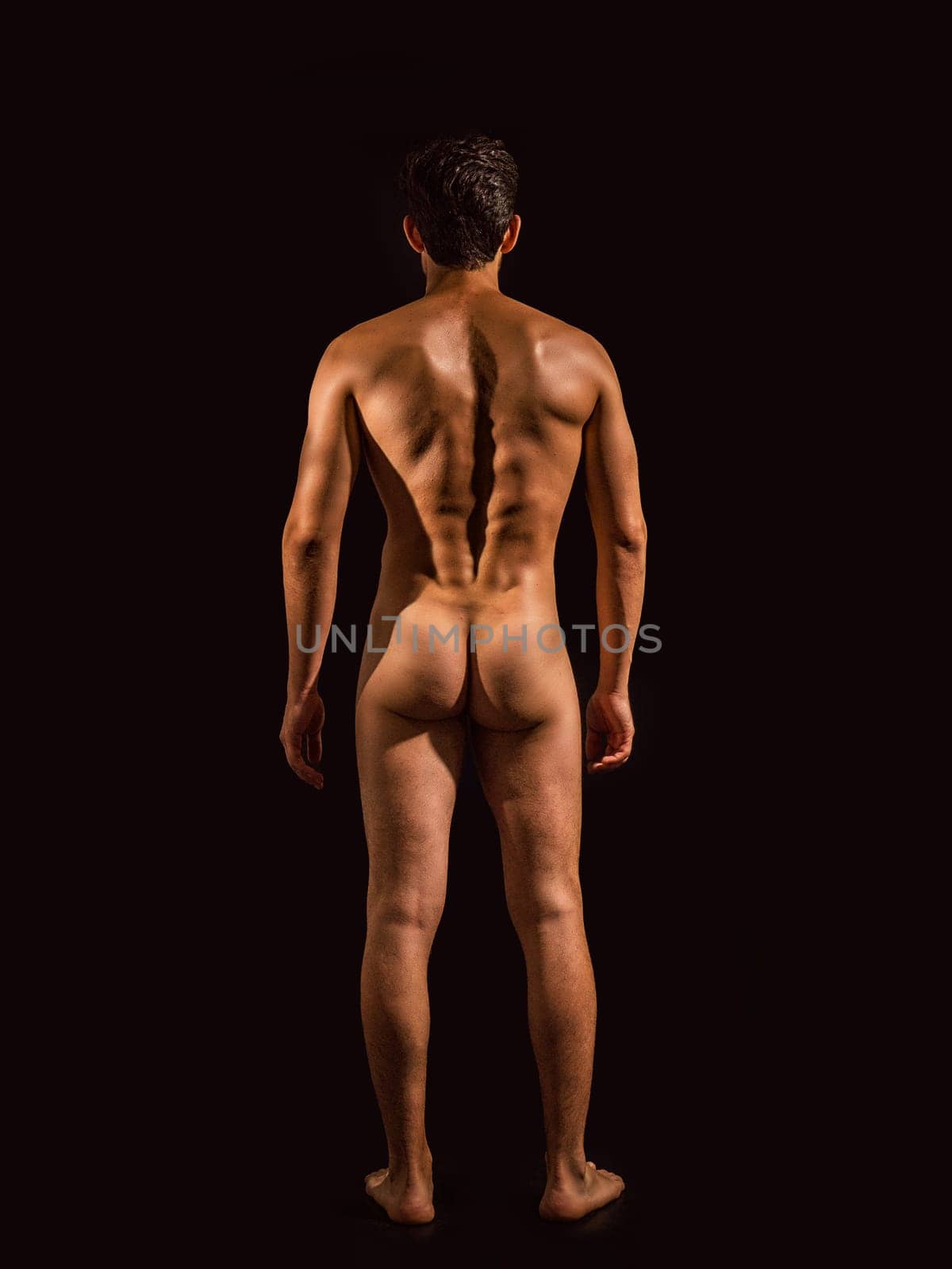 Totally naked athletic handsome young man, seen from the back, showing buttocks, looking straight in a muscular tension pose, on dark background in studio. Portrait of naked handsome young man with languishing look covering crotch with a hand, black background