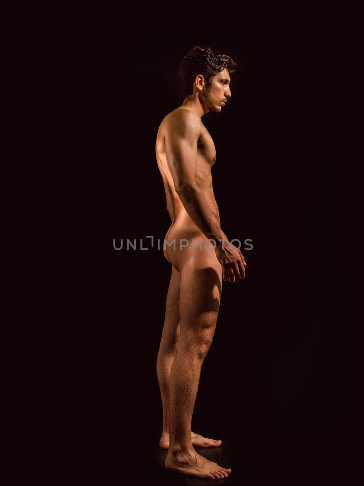 Totally naked athletic handsome young man, seen from the back, showing buttocks, looking straight in a muscular tension pose, on dark background in studio. Portrait of naked handsome young man with languishing look covering crotch with a hand, black background