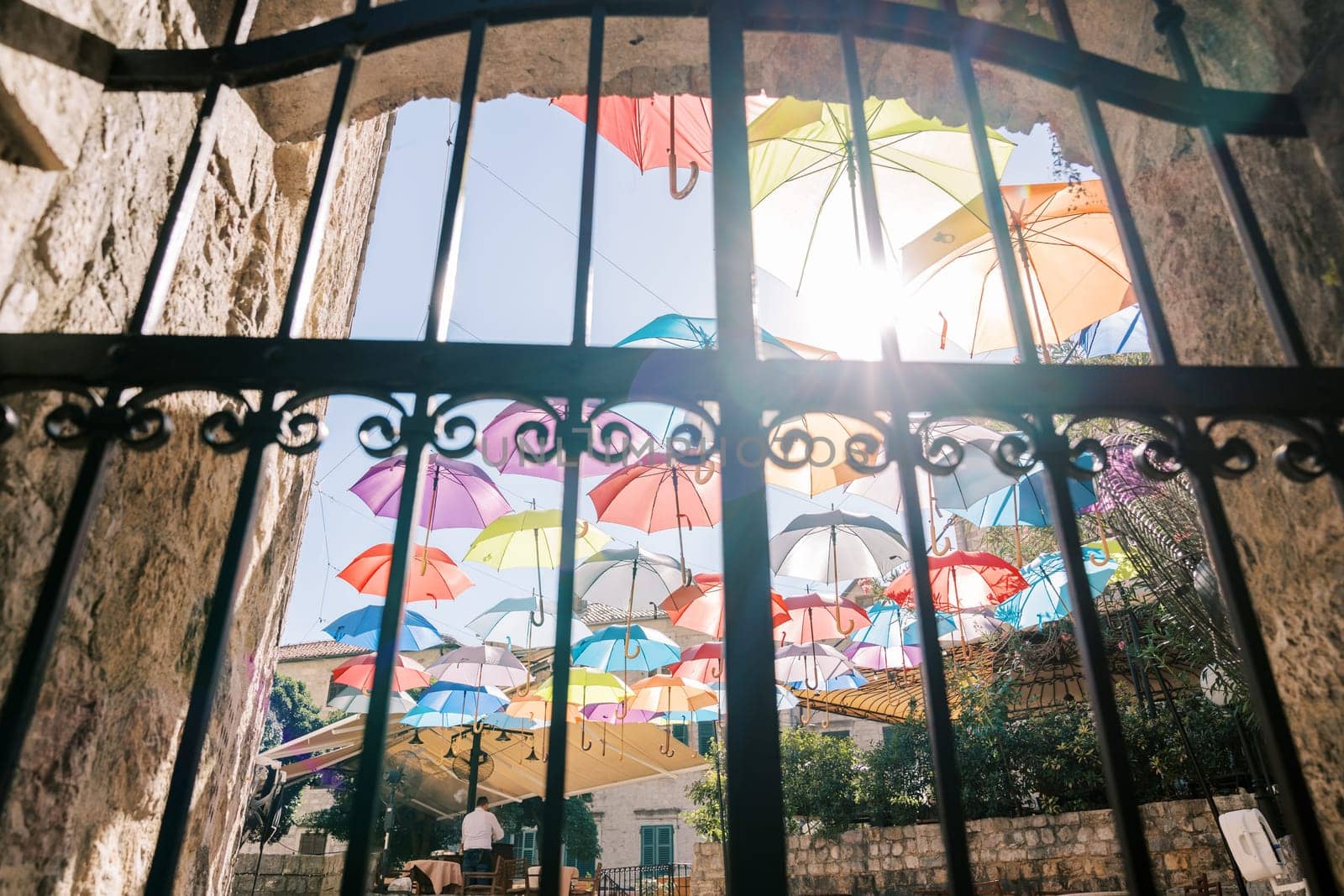 View through a wrought-iron fence to colorful umbrellas hanging above the street of an ancient town by Nadtochiy