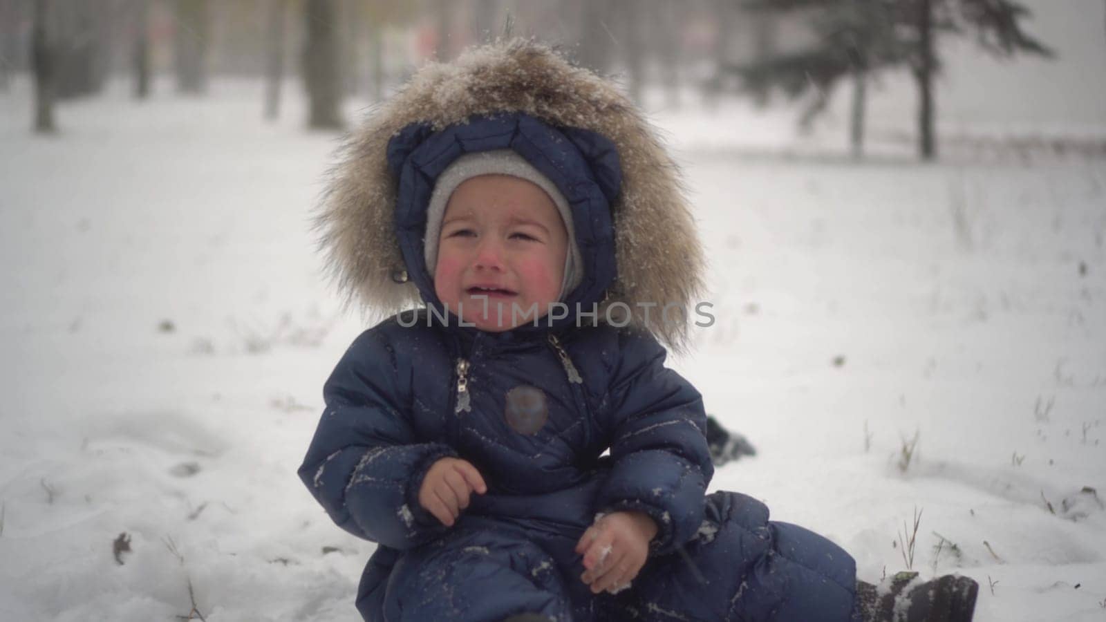 A small child sits in the snow in the park and cries from the cold. The boy in the down overalls had cold hands. 4k