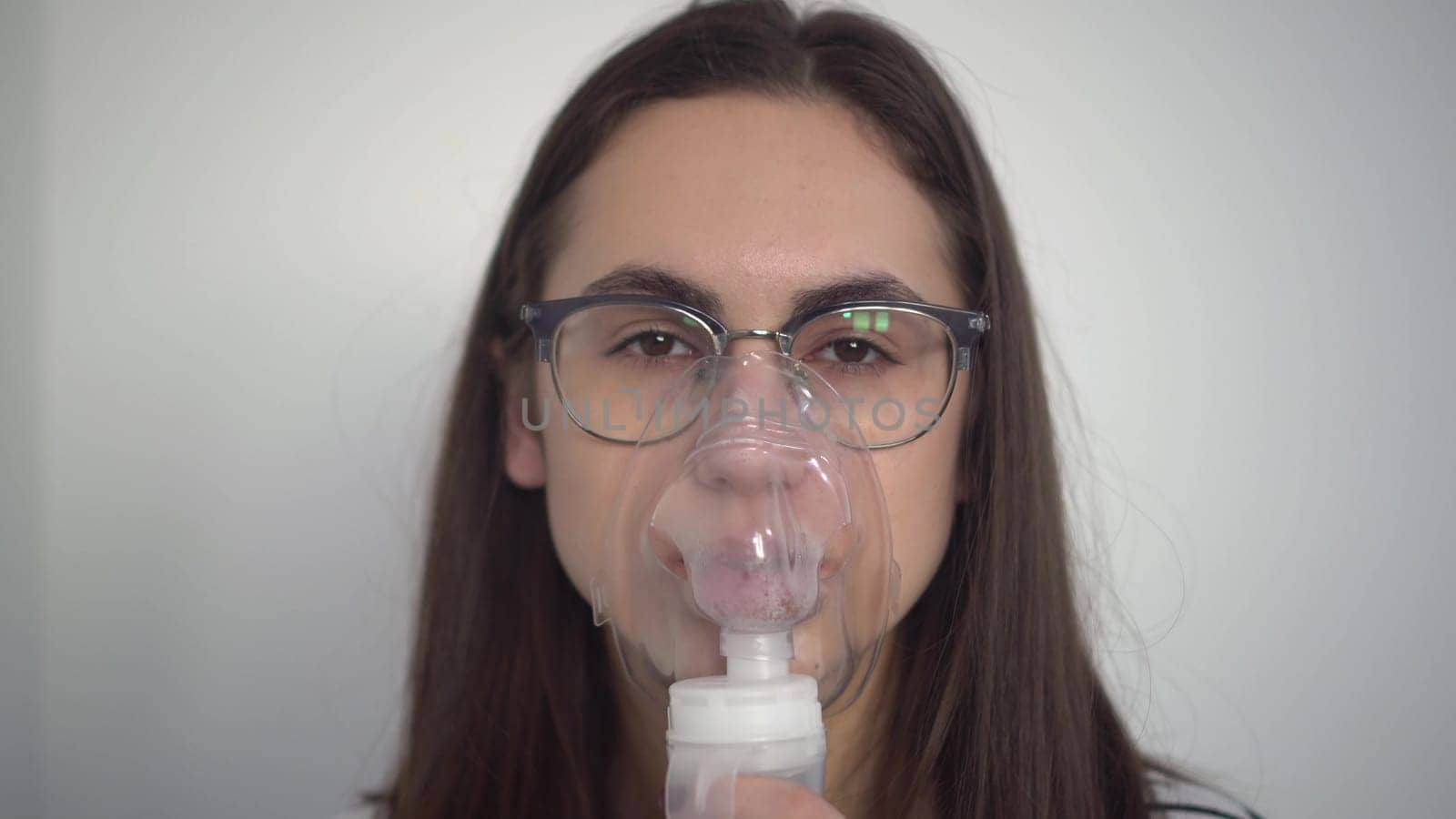 A young woman breathes through an inhaler mask closeup. A girl in glasses with an oxygen mask is being treated for a respiratory infection. by Puzankov