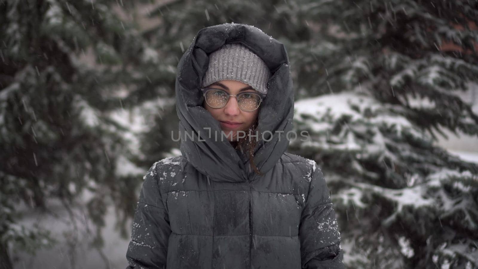 A young woman stands against a background of fir trees under heavy snowfall in winter. A girl in glasses and a down jacket with a hood stands and looks at the camera. Closeup. 4k