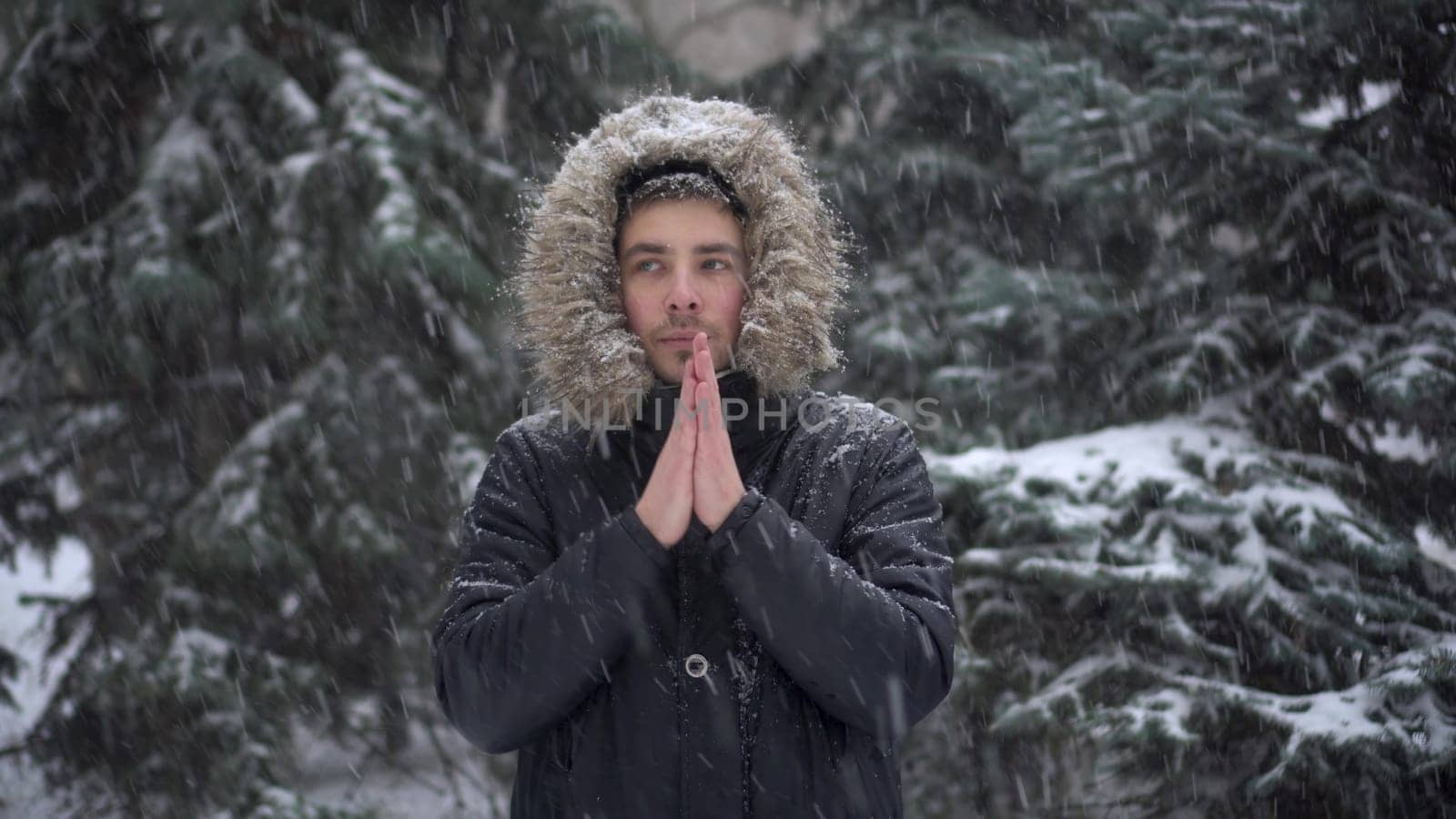 A young man stands against a background of fir trees under heavy snowfall and rubs his hands against the cold. A man in a down jacket with a hood stands and looks at the camera. 4k