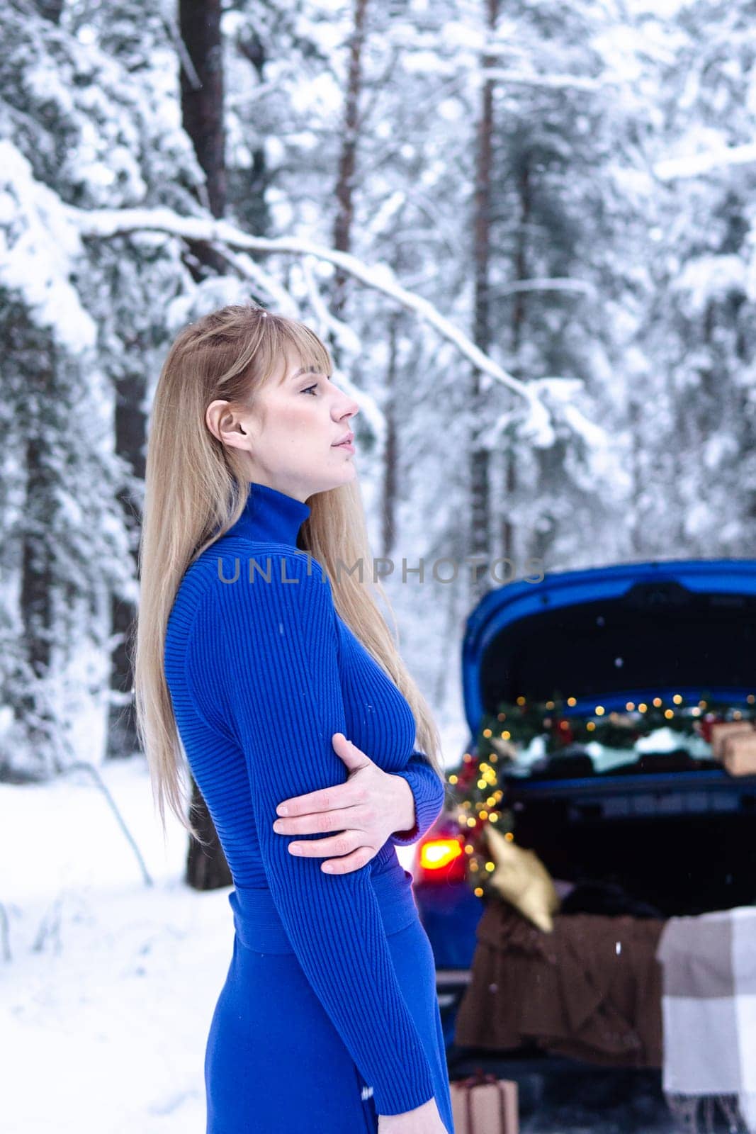 Woman in winter snowy forest in blue dress next to blue car decorated with Christmas decor. Christmas and winter holidays concept