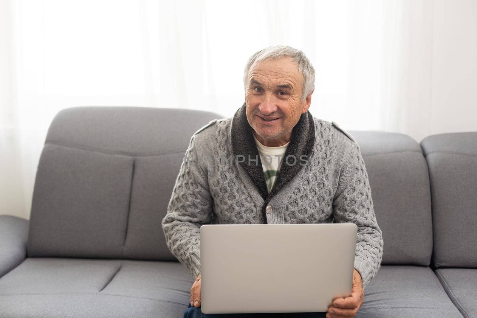 Older man sitting on sofa, smiling at computer screen at home. High quality photo