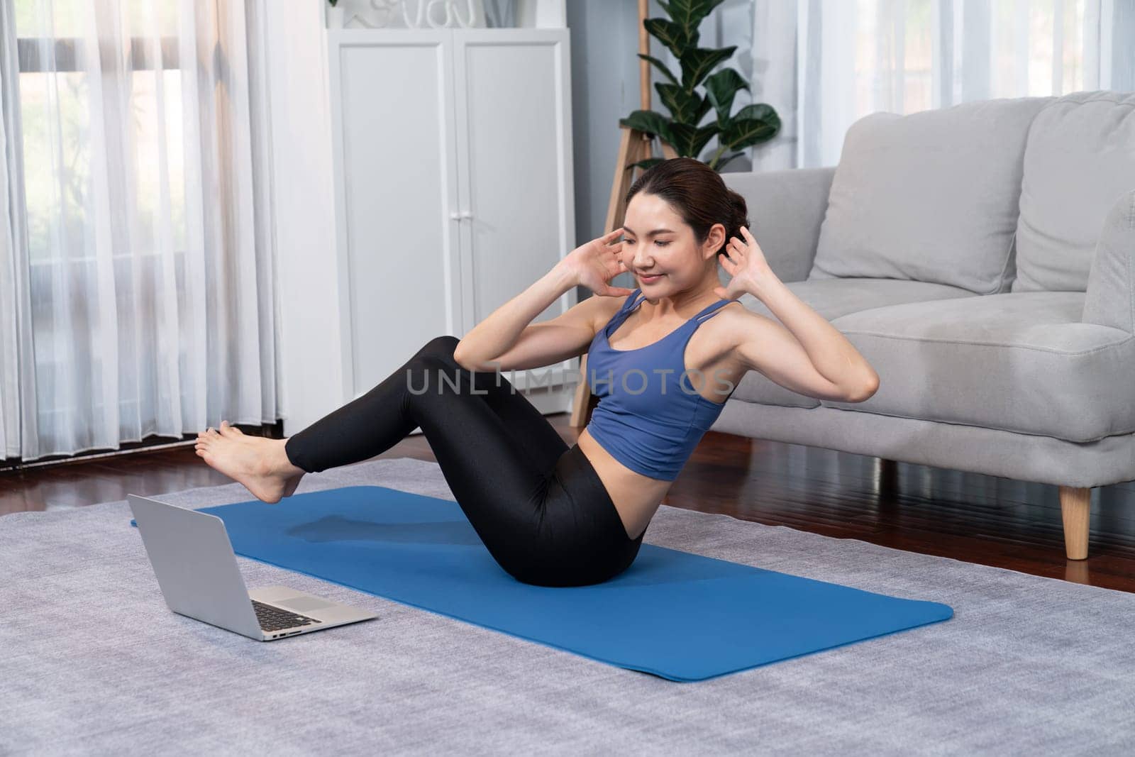 Asian woman in sportswear doing crunch on exercising mat as home workout training routine. Attractive girl engage in her pursuit of healthy lifestyle with online exercise training video. Vigorous