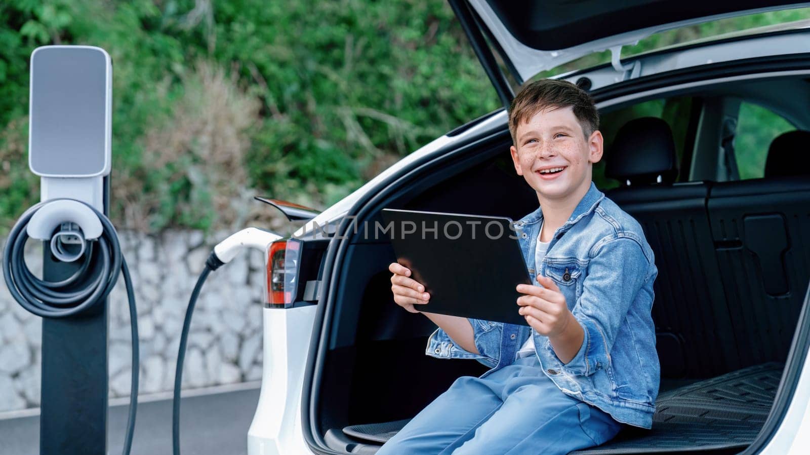 Little boy sitting on car trunk, using tablet while recharging eco-friendly car from EV charging station. EV car road trip travel as alternative vehicle using sustainable energy concept. Perpetual