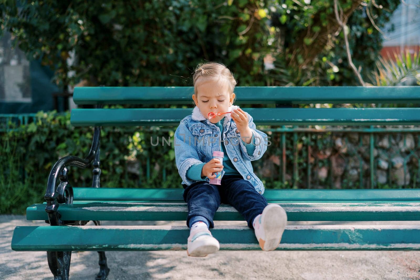 Little girl sits on a bench and blows soap bubbles by Nadtochiy