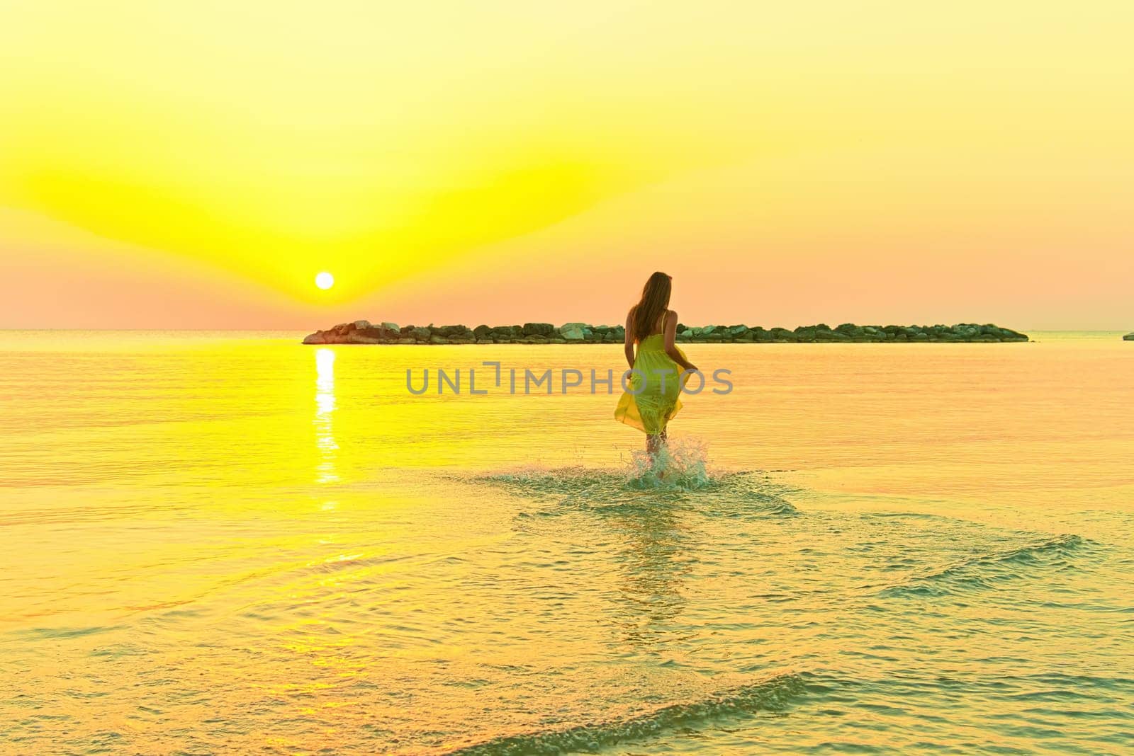 A young woman in a bright yellow dress meets the sunrise, joyfully runs through the sea water