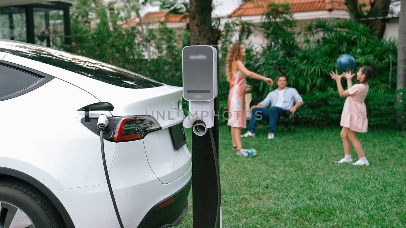 Electric vehicle recharge from home charging station on background of happy and playful family playing together. EV car using alternative and sustainable energy for better future Synchronos