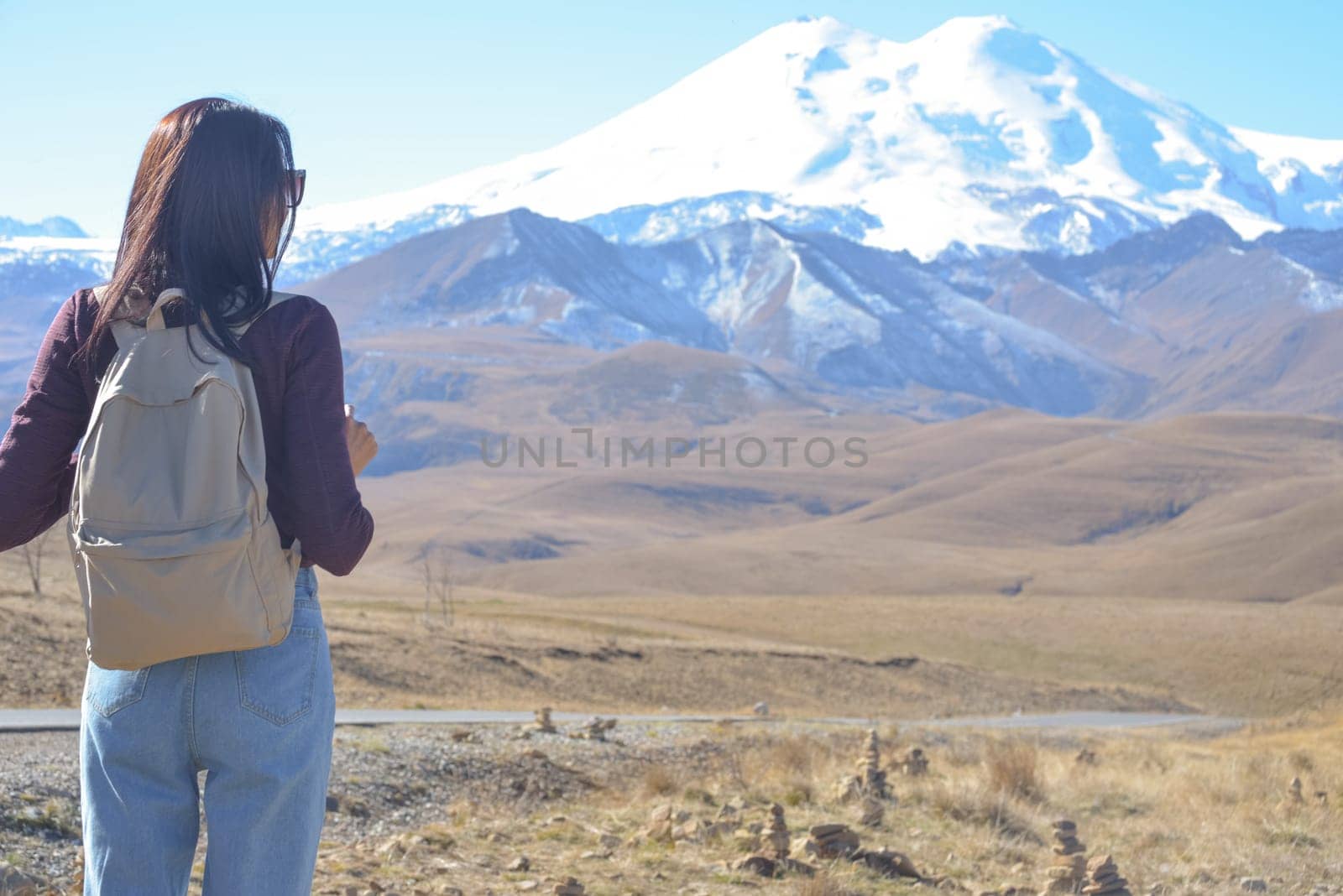 A female traveler with a backpack on her back admires the snow-covered Elbrus on a clear autumn day. View of Elbrus, North Caucasus, Russia.