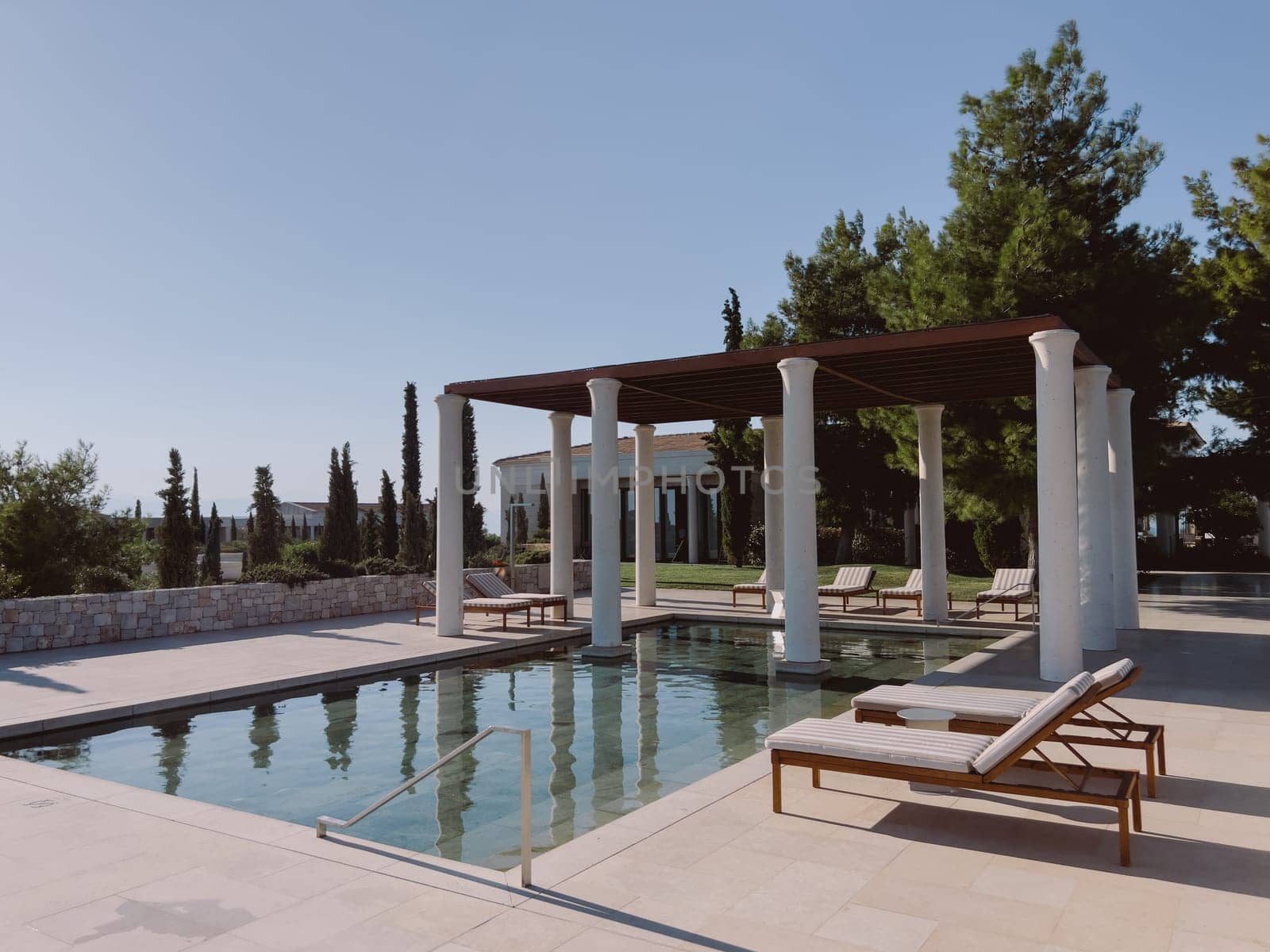 Pergola with columns near the pool with sun loungers surrounded by trees. Hotel Amanzoe, Greece. High quality photo