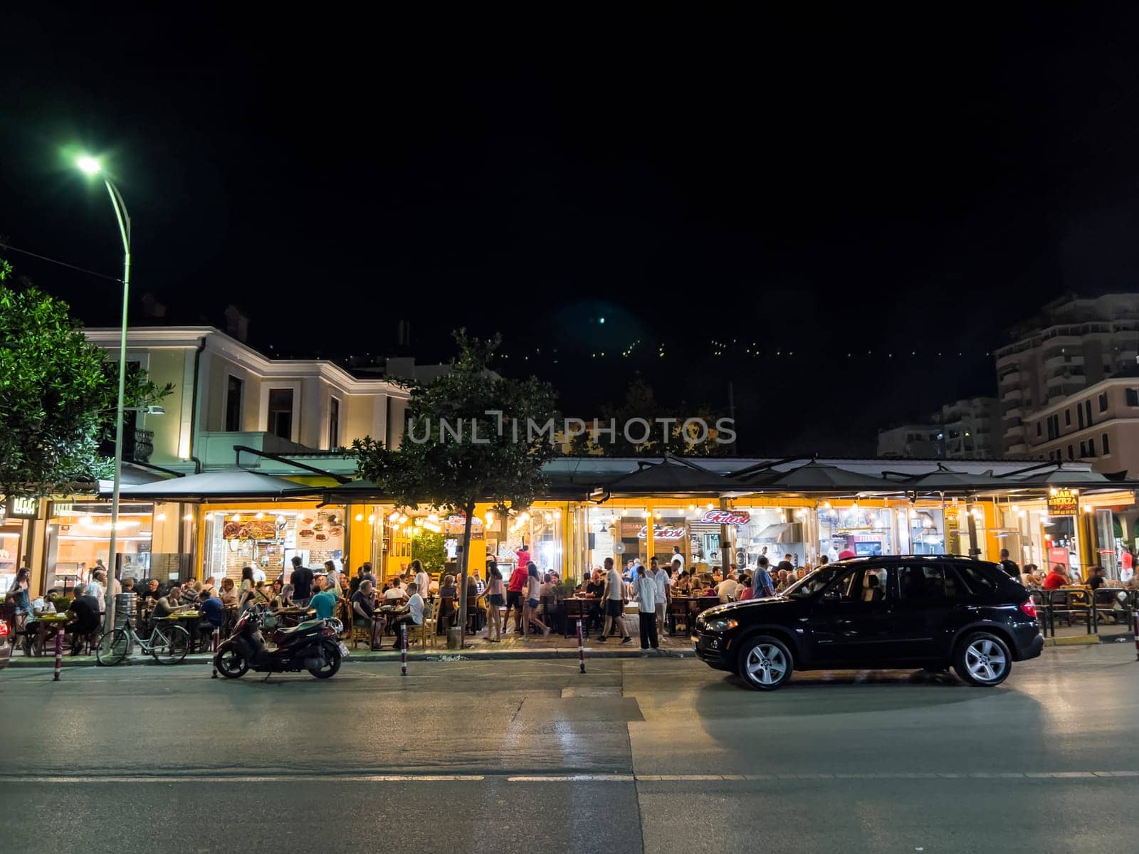 Budva, Montenegro - 05 august 2023: People sit at the tables of a street cafe lit by lamps near the road by Nadtochiy